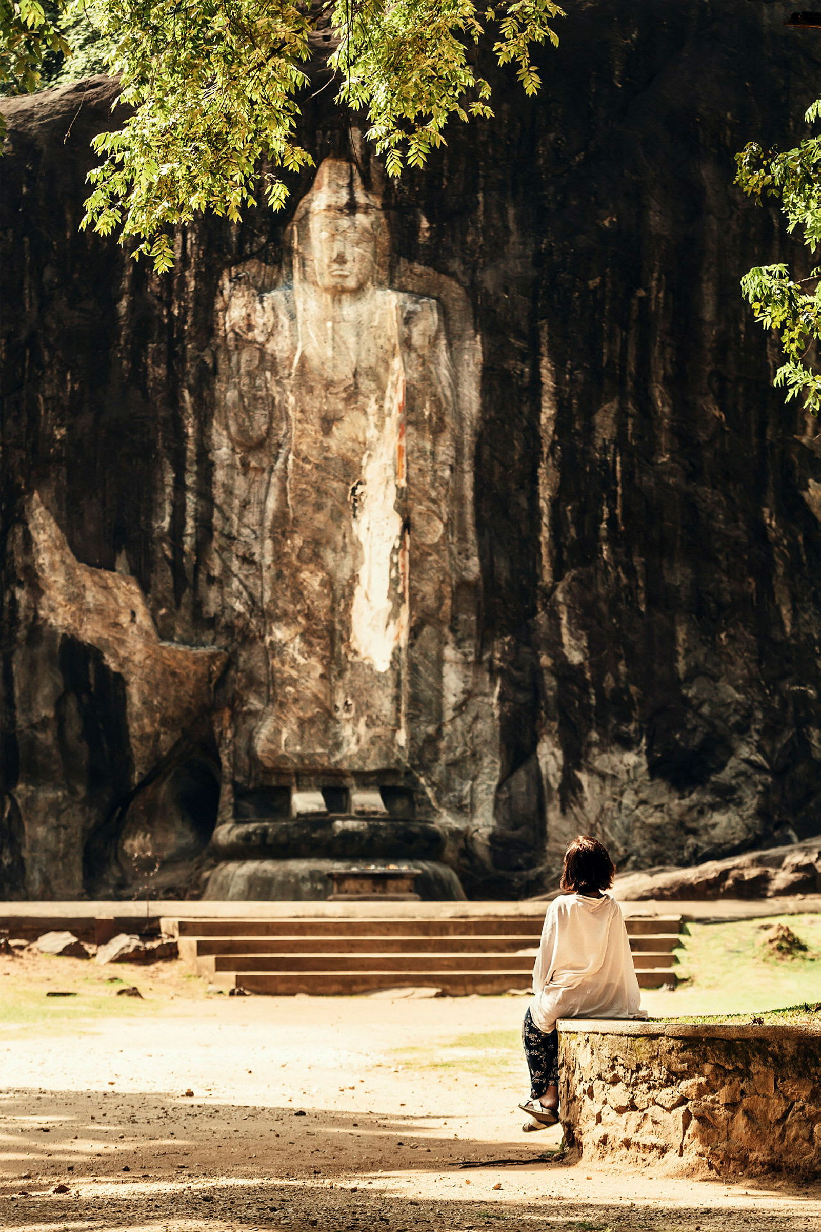 En person sitter på en stenmur och tittar mot en stor buddhistisk staty ristad i en bergvägg. (AI-genererad text)