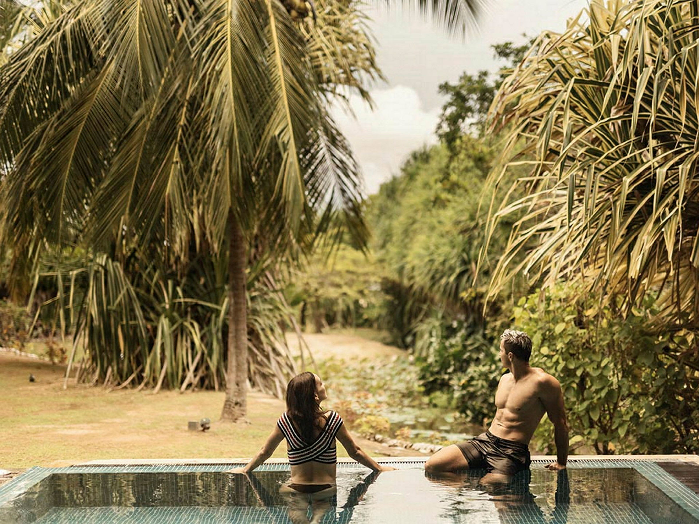 Två personer sitter på kanten av en pool med frodig vegetation och palmer i bakgrunden. (AI-genererad text)