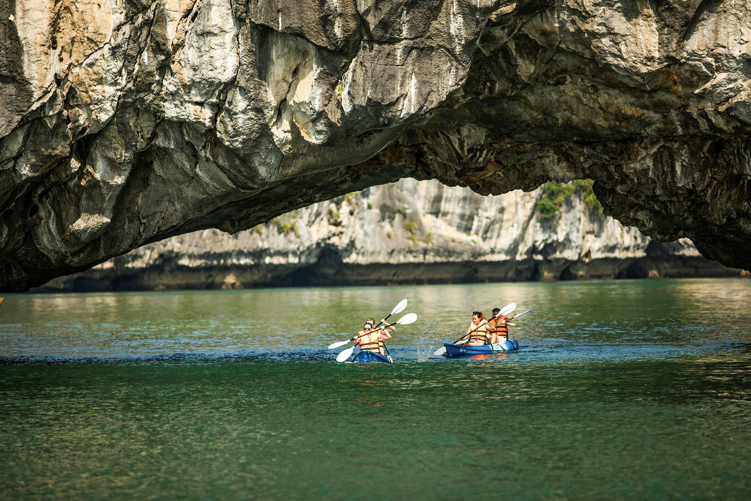 Personer paddlar kajak under en naturlig klippformation över vattnet. (AI-genererad text)
