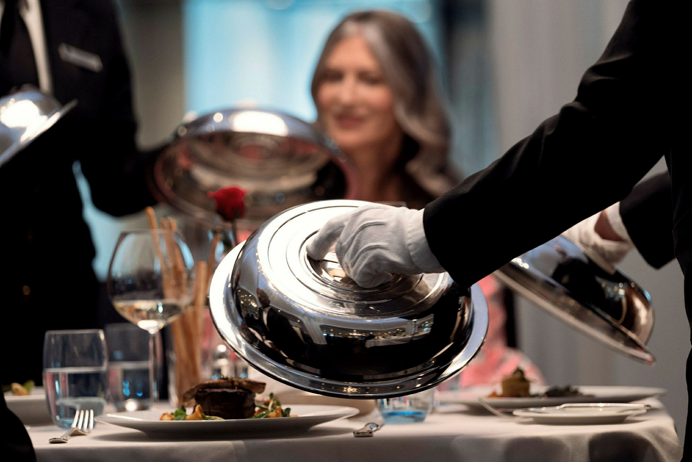 Servitörer lyfter på serveringsklockor vid ett väldukat bord. (AI-genererad text)