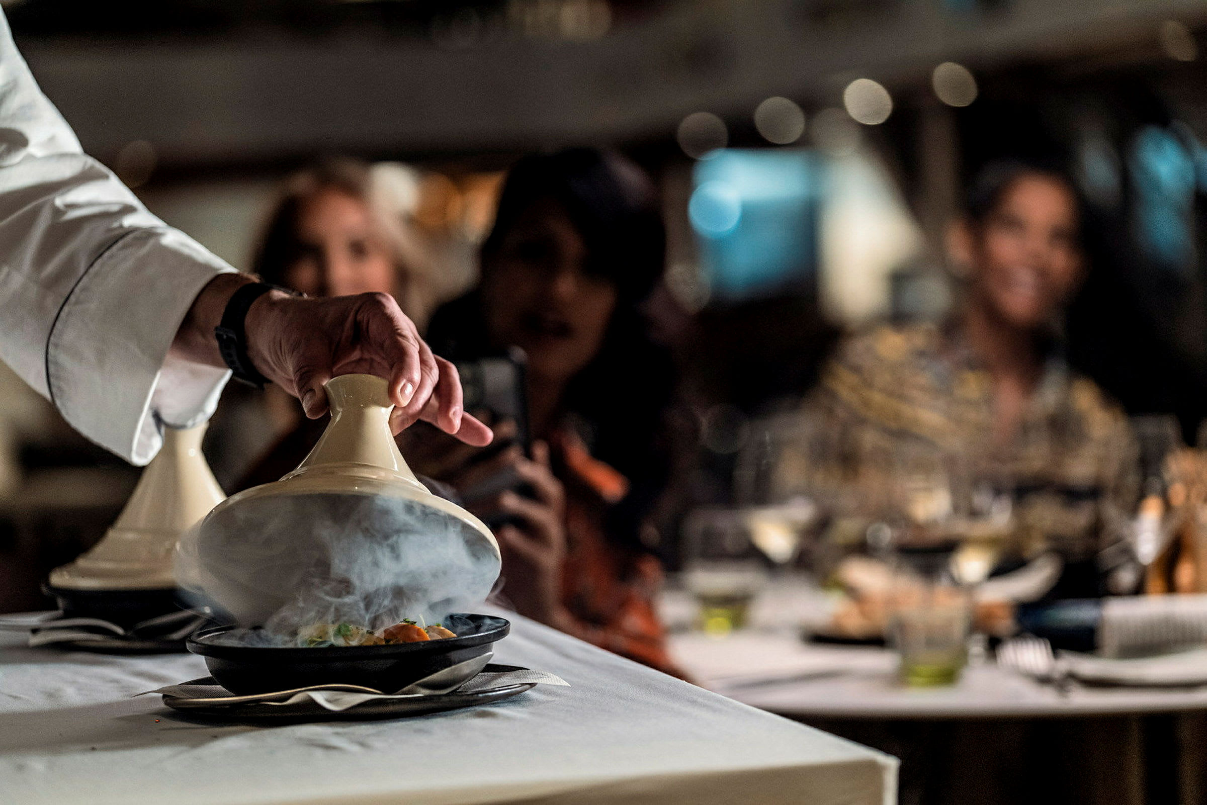 En hand lyfter locket på en tagine på ett bord i en restaurang. (AI-genererad text)