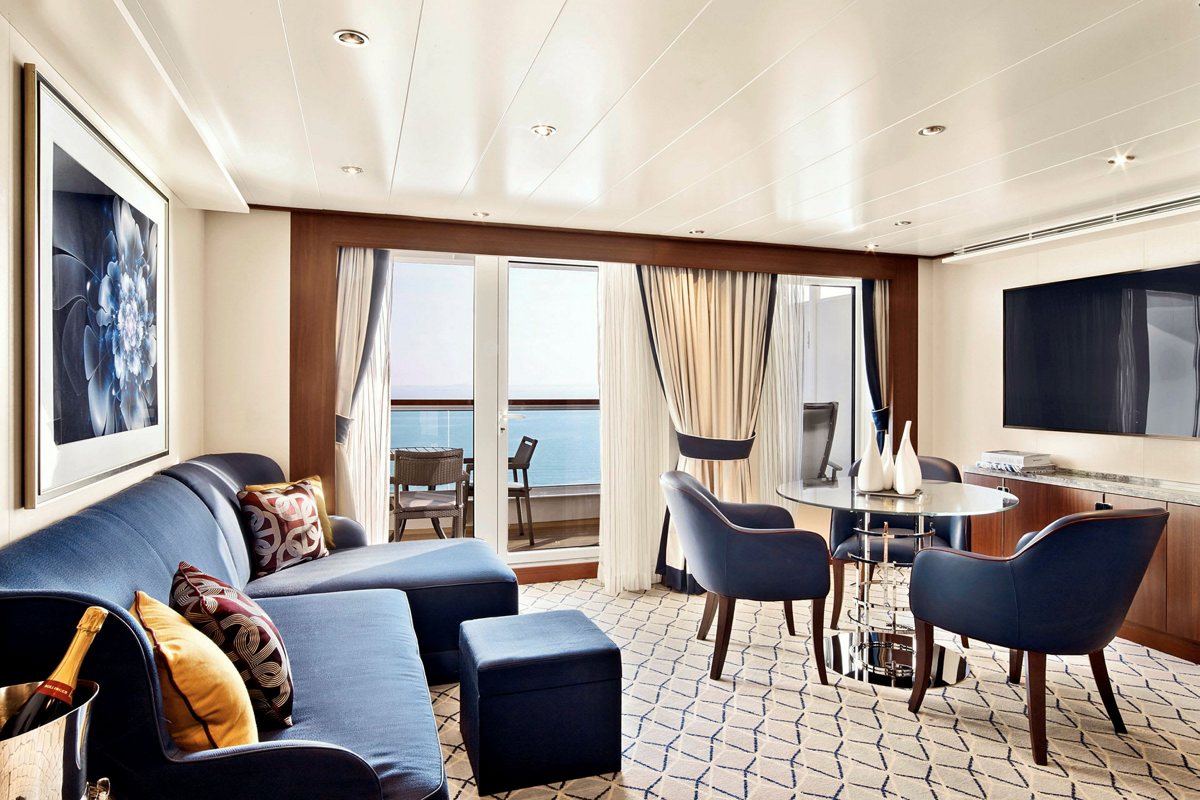 Ett elegant vardagsrum med blå möbler, ett bord och stora fönster mot en balkong med utsikt över havet. (AI-genererad text)