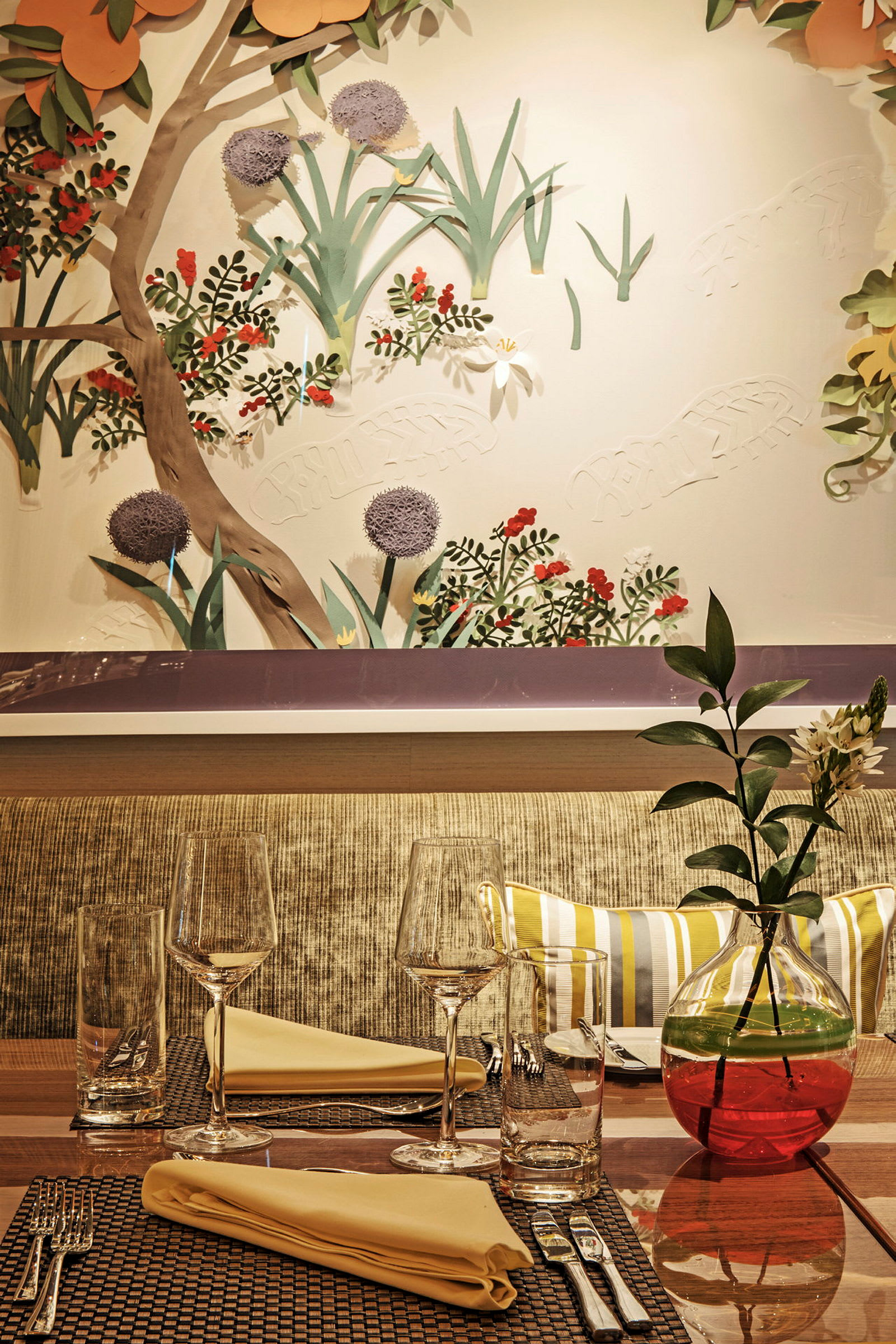Ett väl dukat bord med glas och servetter, samt en dekorativ vas med blommor framför en vägg med naturinspirerad konst. (AI-genererad text)