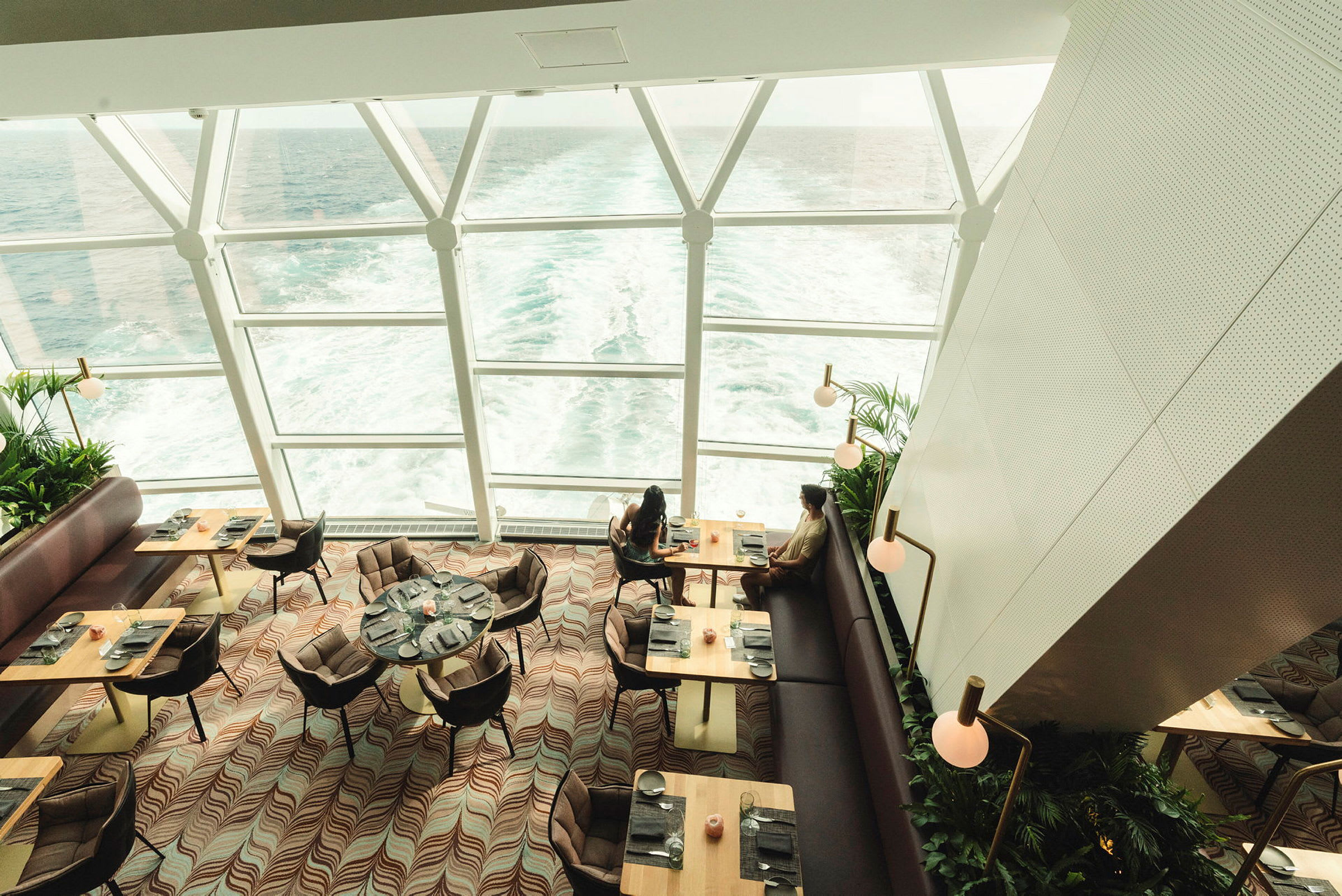 Interiör av en restaurang med stora fönster som visar havet utanför. Syns matbord, stolar och människor som njuter av utsikten. (AI-genererad text)