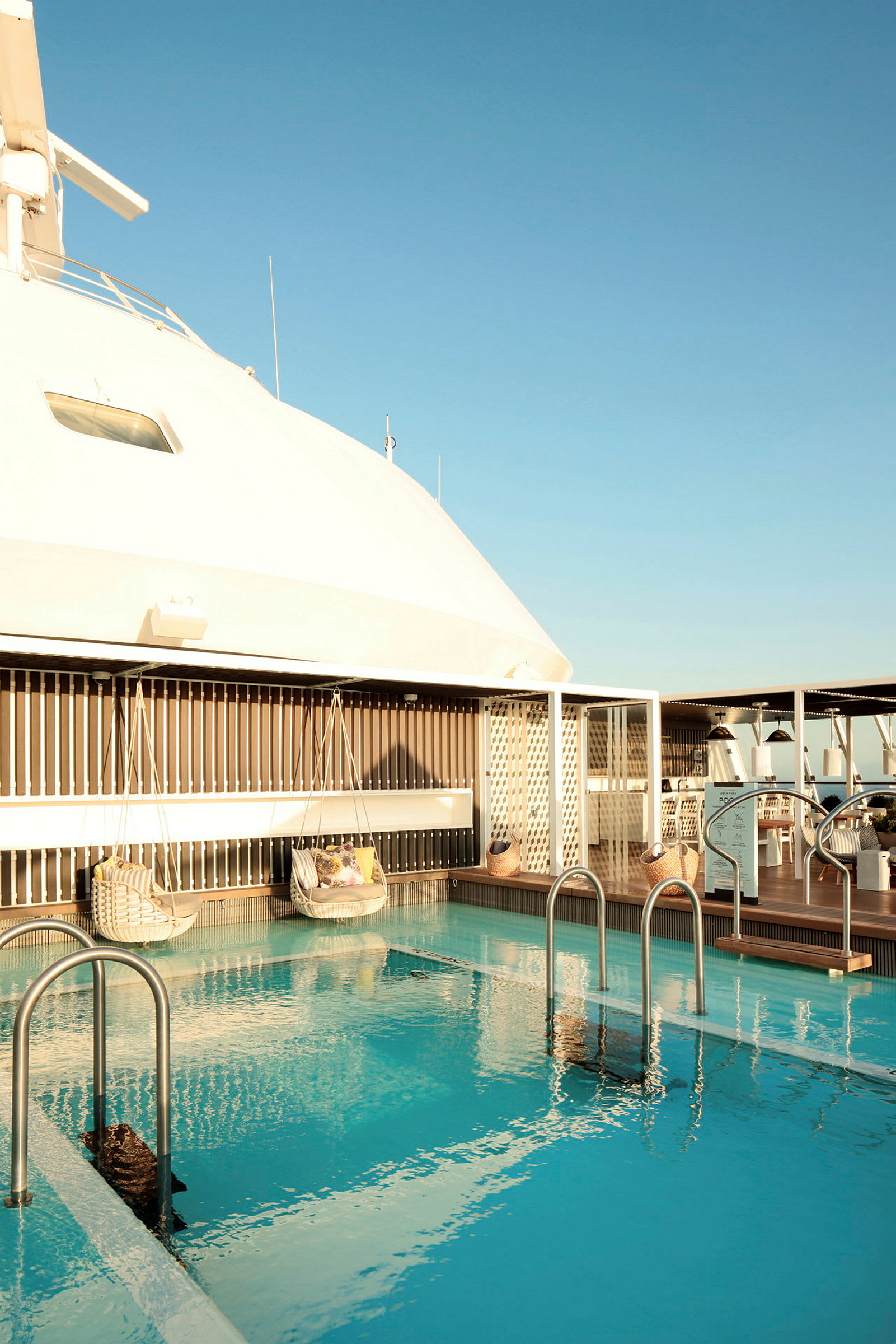 Pool med solstolar och hängande sittplatser på ett fartygsdäck under klar himmel. (AI-genererad text)