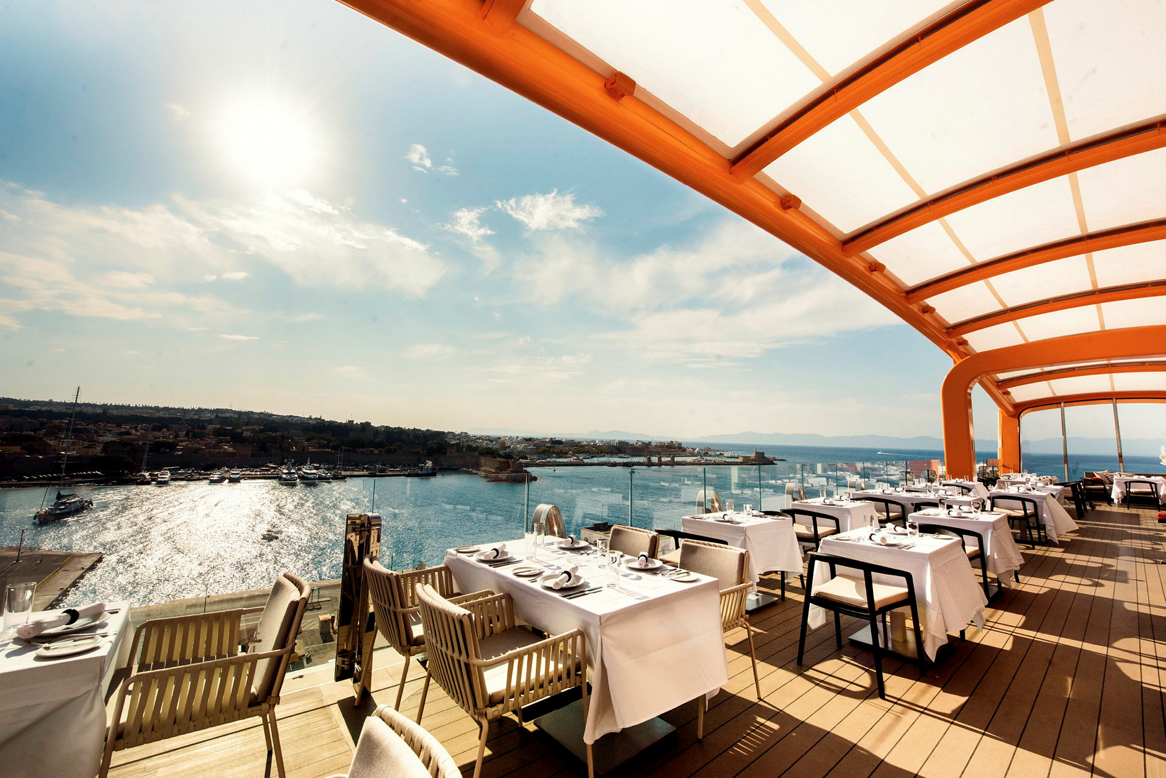 Bild av en restaurang med uteservering under ett täckande tak, med havet och några båtar i bakgrunden. (AI-genererad text)