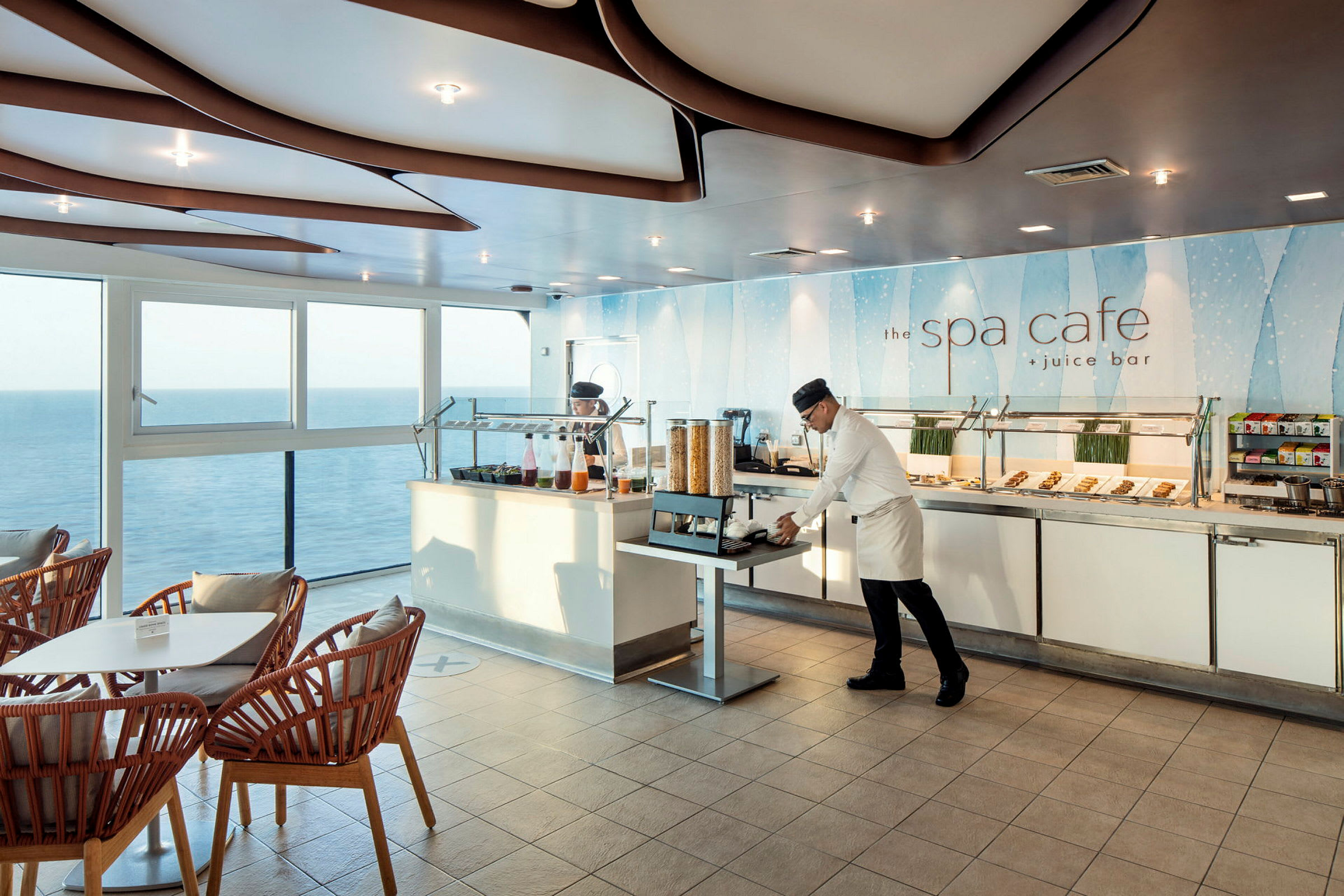 Ett kafé med stora fönster som visar utsikt över havet, med sittplatser och en buffé fylld med mat och dryck. (AI-genererad text)