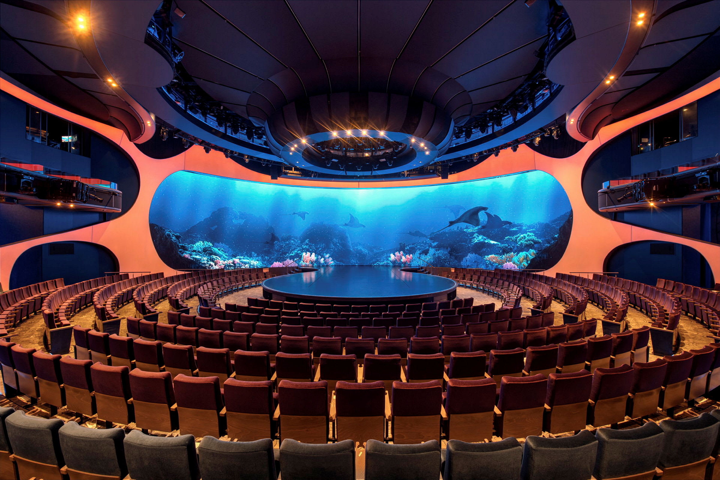 Ett auditorium med en rund scen och rader av publikstolar. Bakgrunden består av en stor skärm som visar ett undervattensmotiv. (AI-genererad text)
