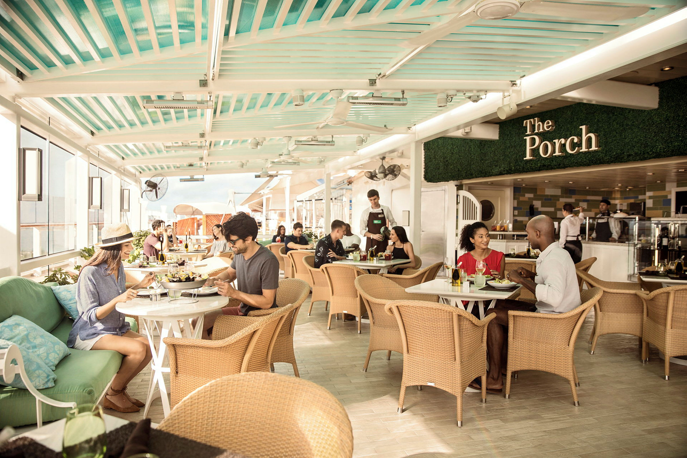 En restaurang med gäster som sitter vid bord och äter och pratar. Inredningen har rottingstolar och ett soltak. (AI-genererad text)
