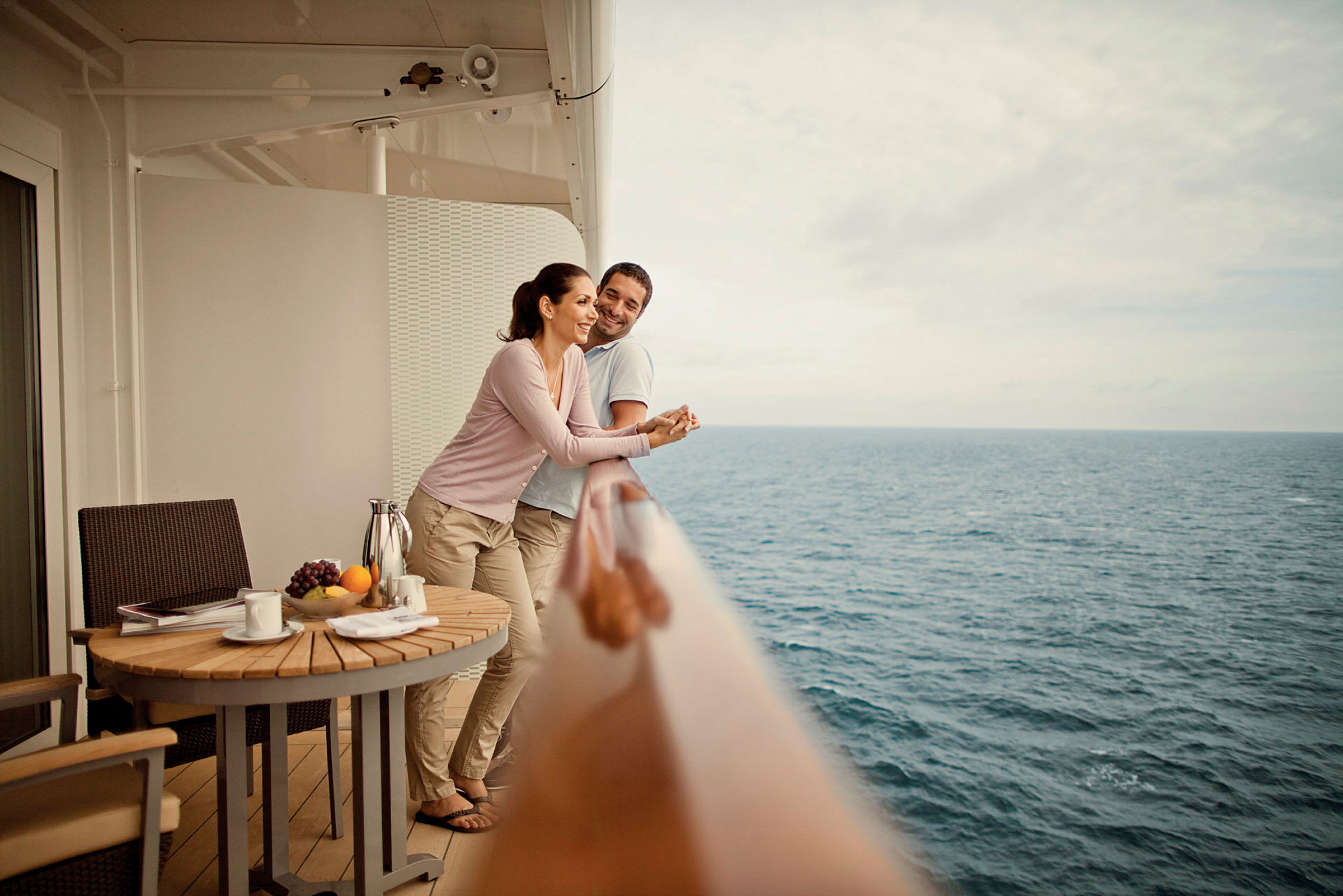 Personer står på en balkong med utsikt över havet. En träbord är placerad med frukter och drycker. (AI-genererad text)