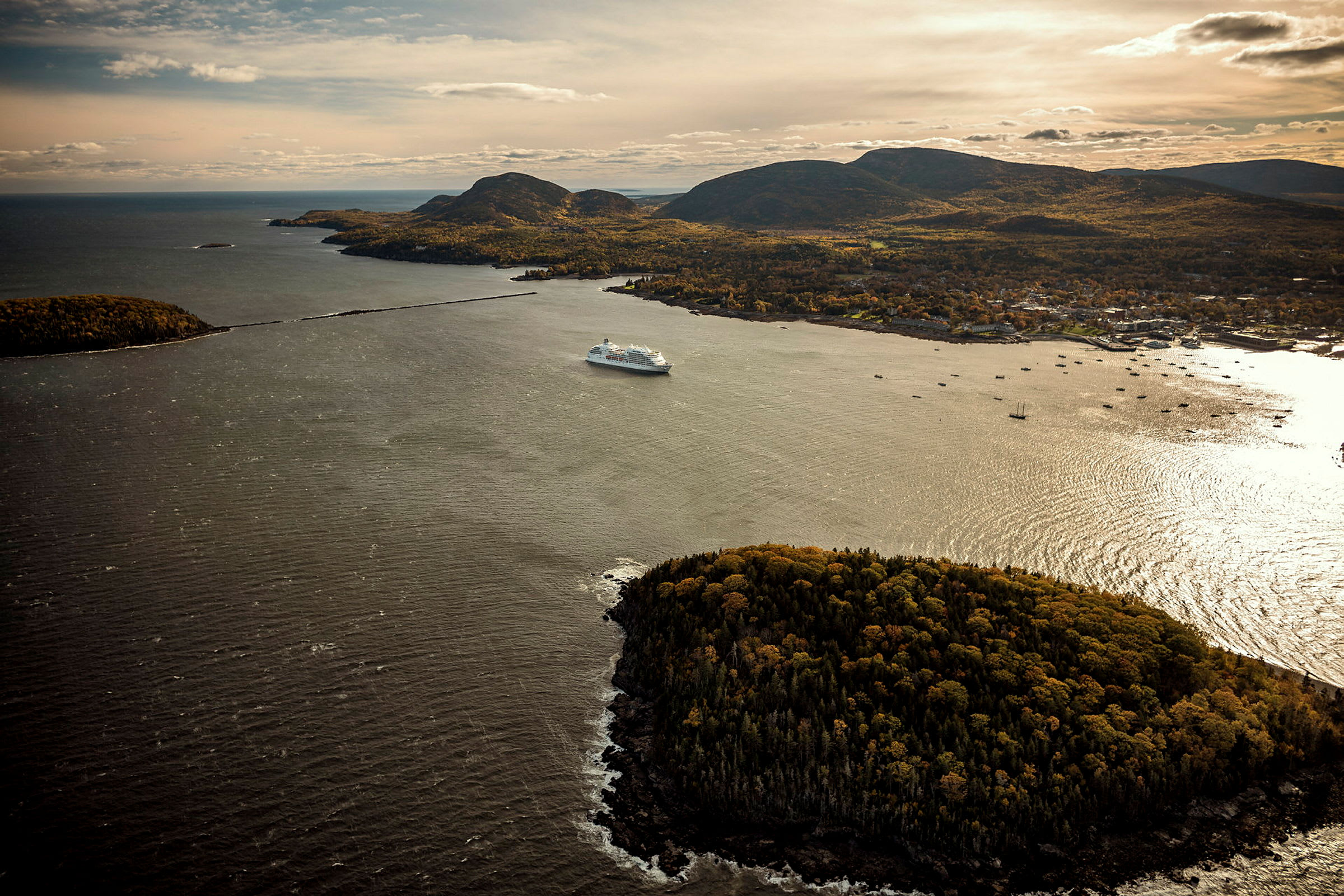 Fågelperspektiv av ett stort kryssningsfartyg vid en kust med skogklädd halvö och berg i bakgrunden. (AI-genererad text)