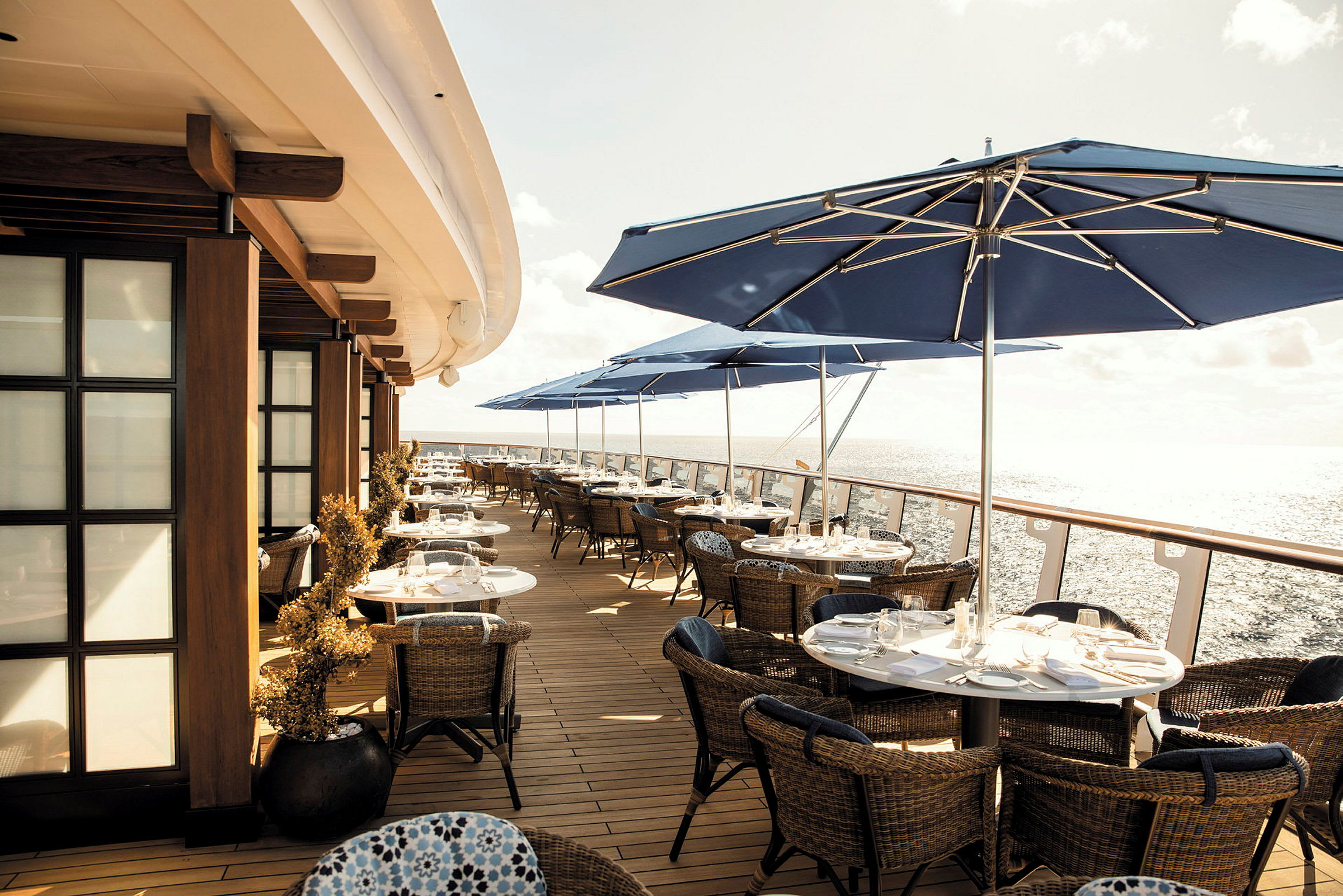 En utomhusrestaurang med bord, stolar och blå parasoller, med utsikt över havet. (AI-genererad text)