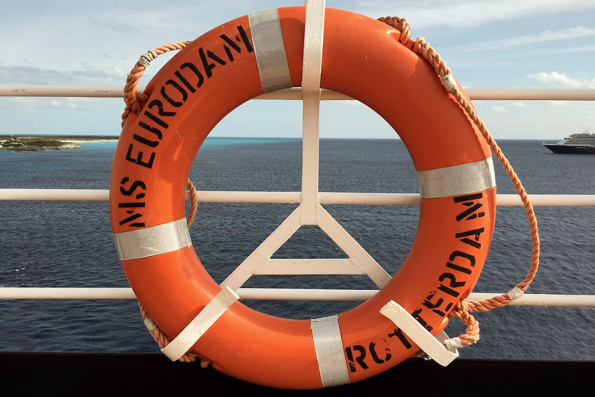 En orange livboj hängande på ett fartygs reling med hav och en ö i bakgrunden. (AI-genererad text)