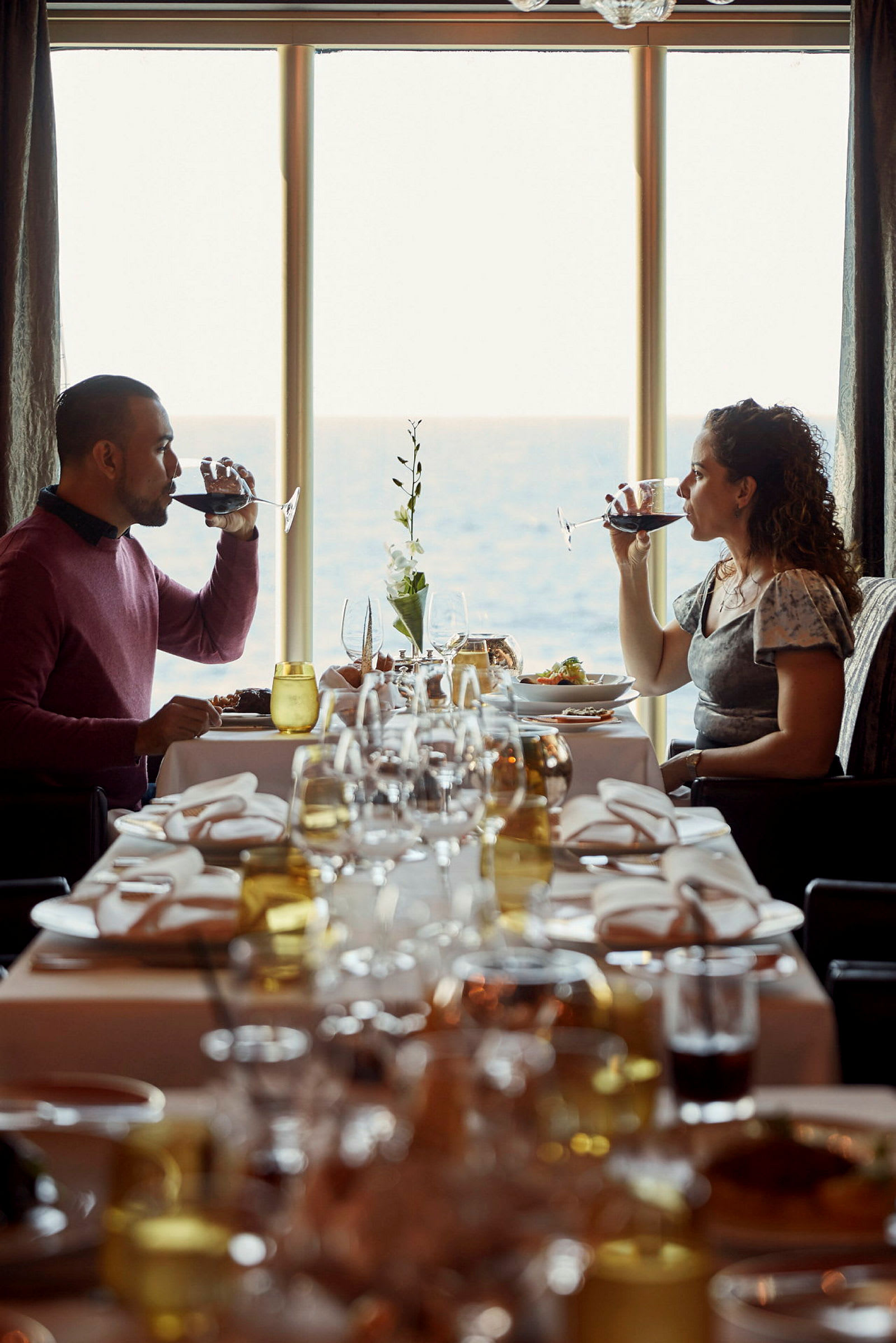 Två personer sitter vid ett matbord med duk och glas, med havet synligt genom ett stort fönster. (AI-genererad text)