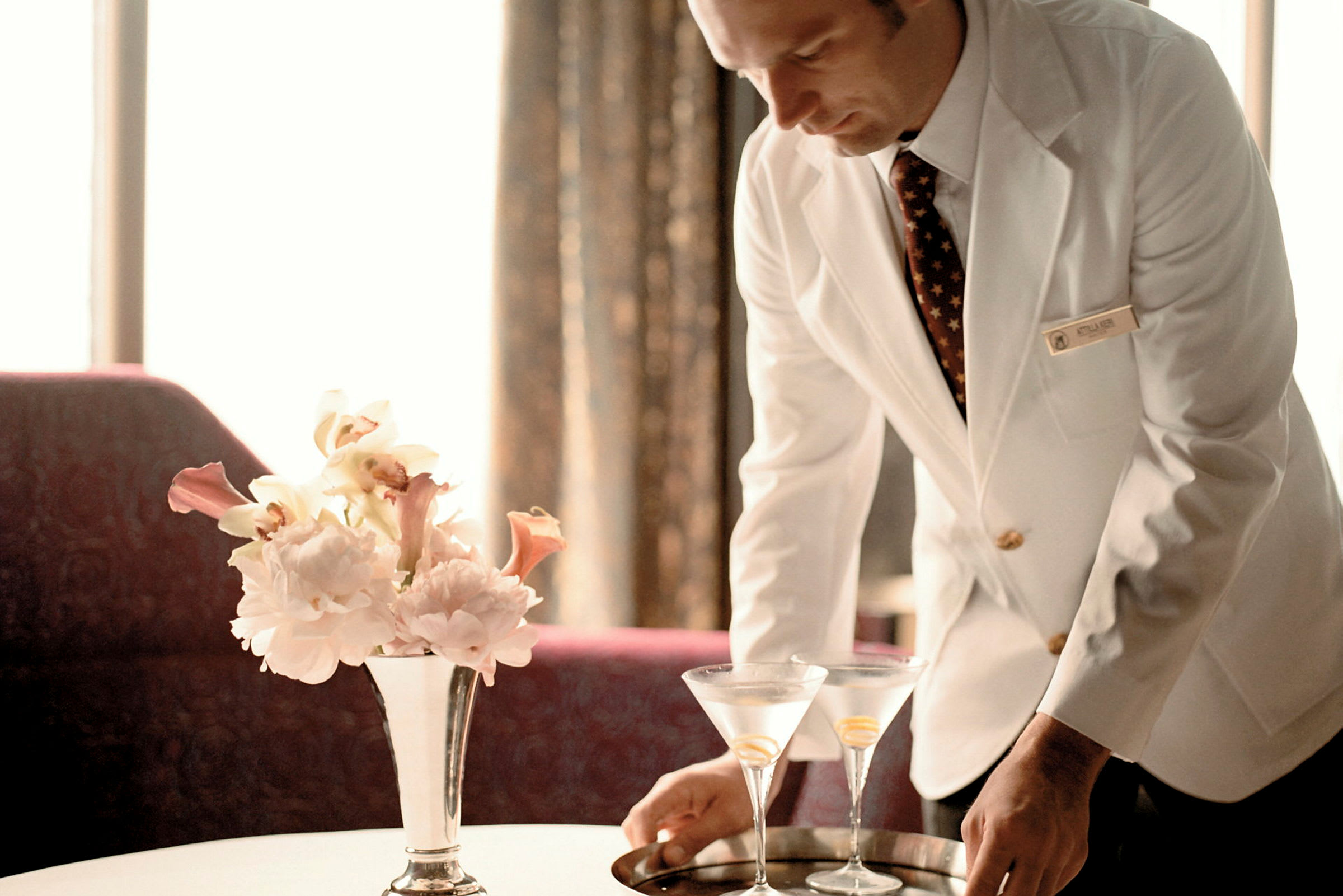 En person i vitt hotellpersonaluniform arrangerar två glas på ett brickbord bredvid en vas med blommor. (AI-genererad text)