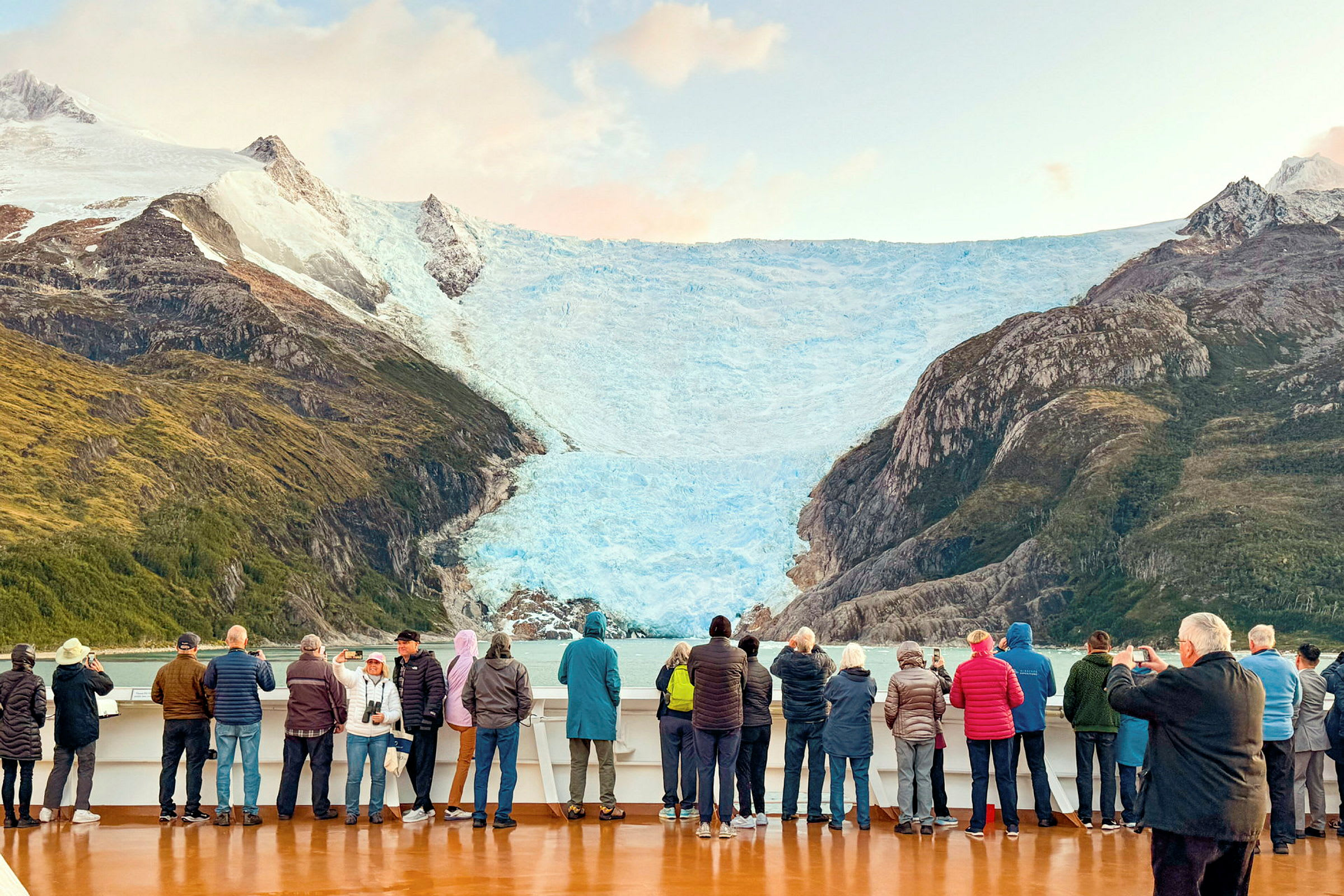 En grupp människor står på ett fartygsdäck och beskådar en stor glaciär med runt omkringliggande berg. (AI-genererad text)