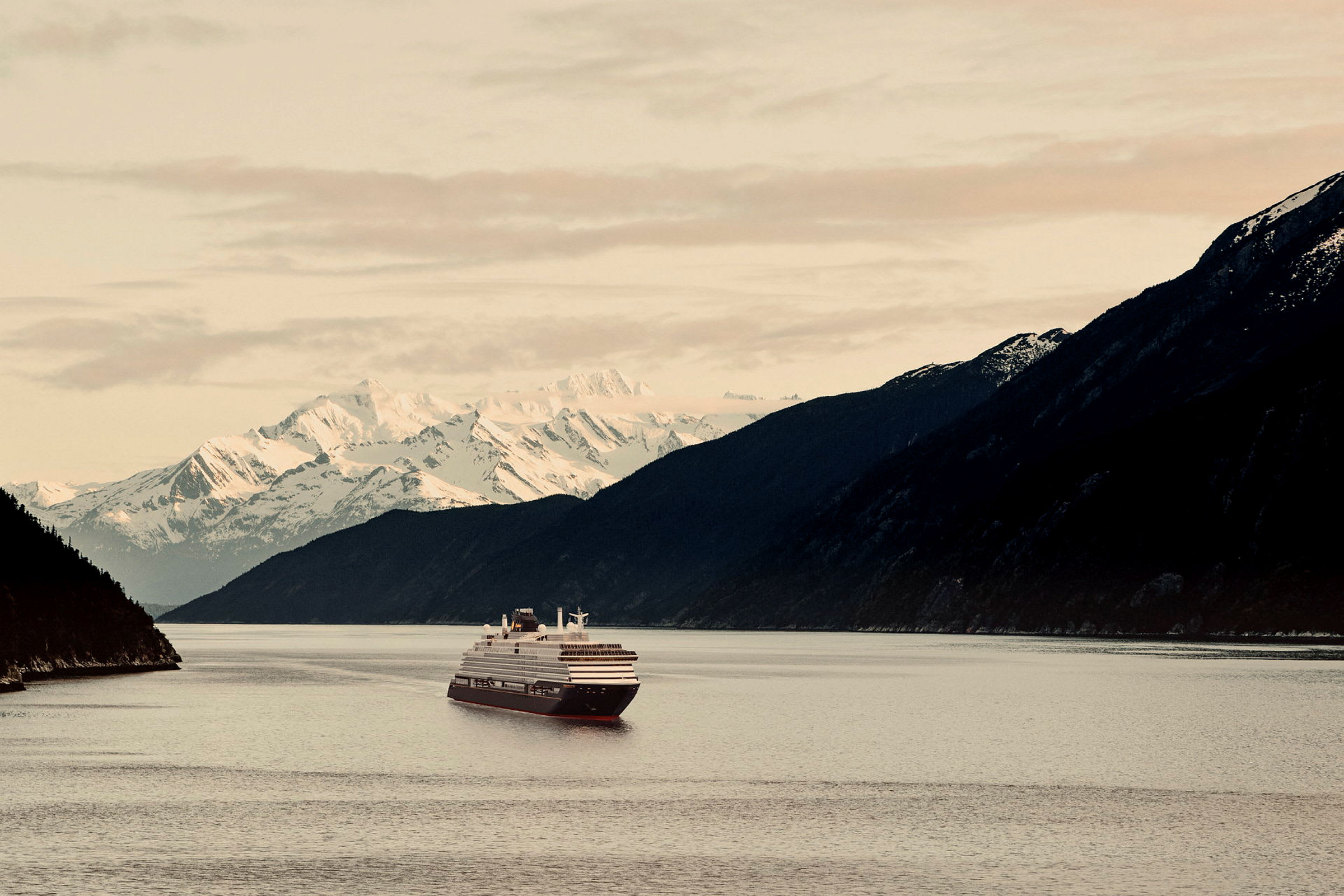 Ett kryssningsfartyg seglar på en bred fjord, omgiven av höga berg och snötäckt landskap. (AI-genererad text)