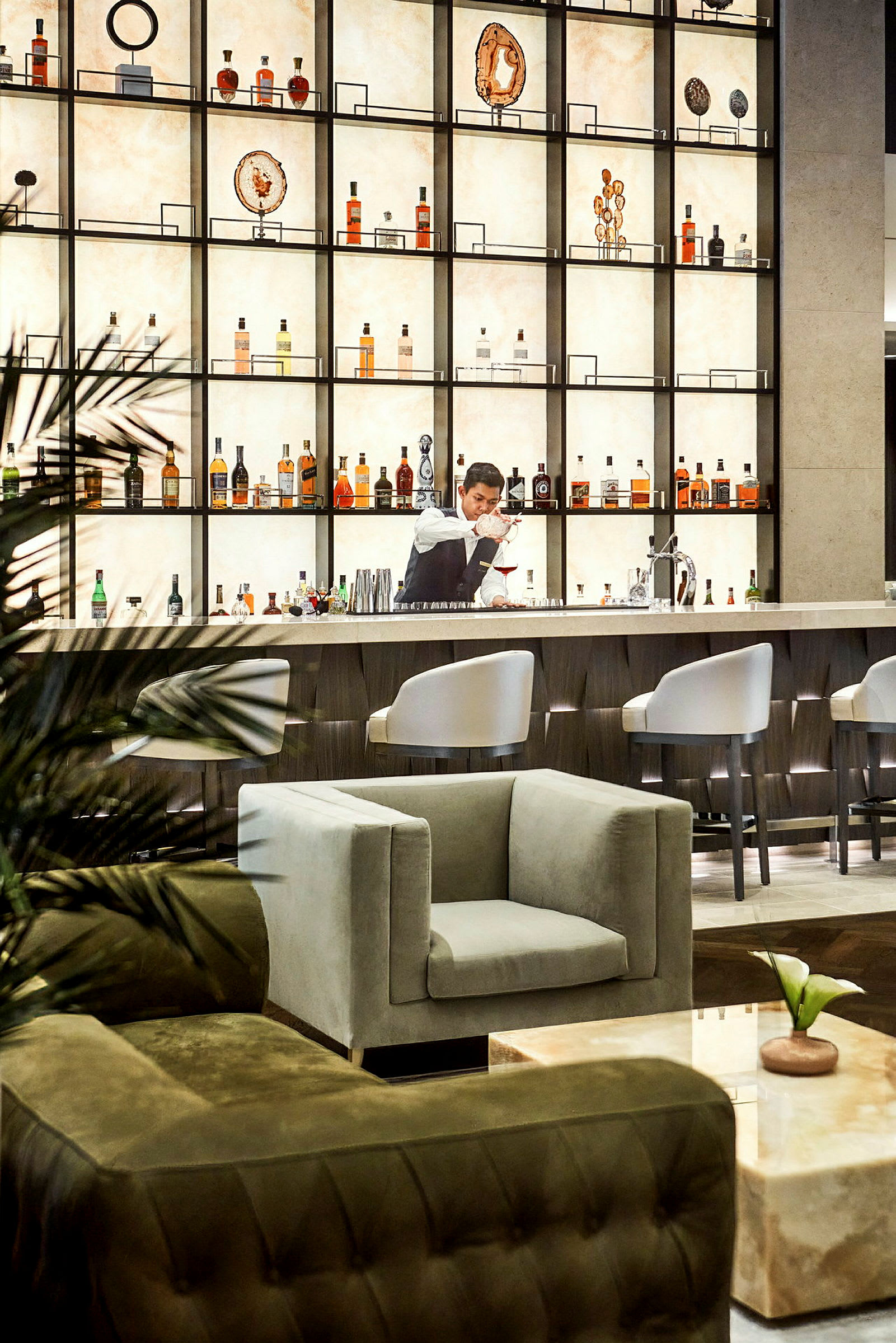En elegant bar med en hylla fylld med flaskor och prydnadsföremål, samt fåtöljer och bord i förgrunden. (AI-genererad text)