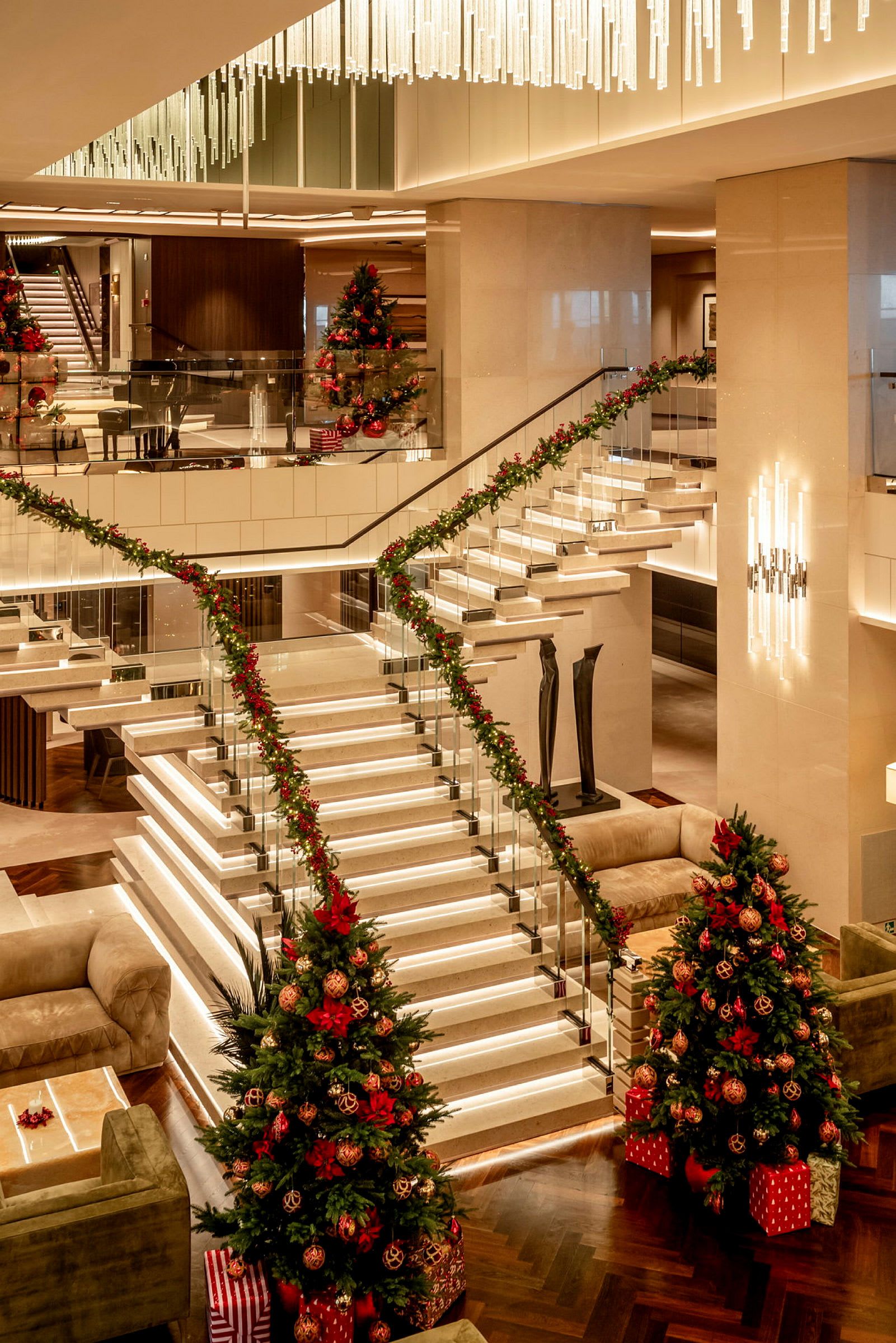 Ett elegant hotelllobby med en centralt placerad trappa dekorerad med julgranspynt och belysta detaljer, omgiven av soffor och julgranar med prydnader och presenter. (AI-genererad text)