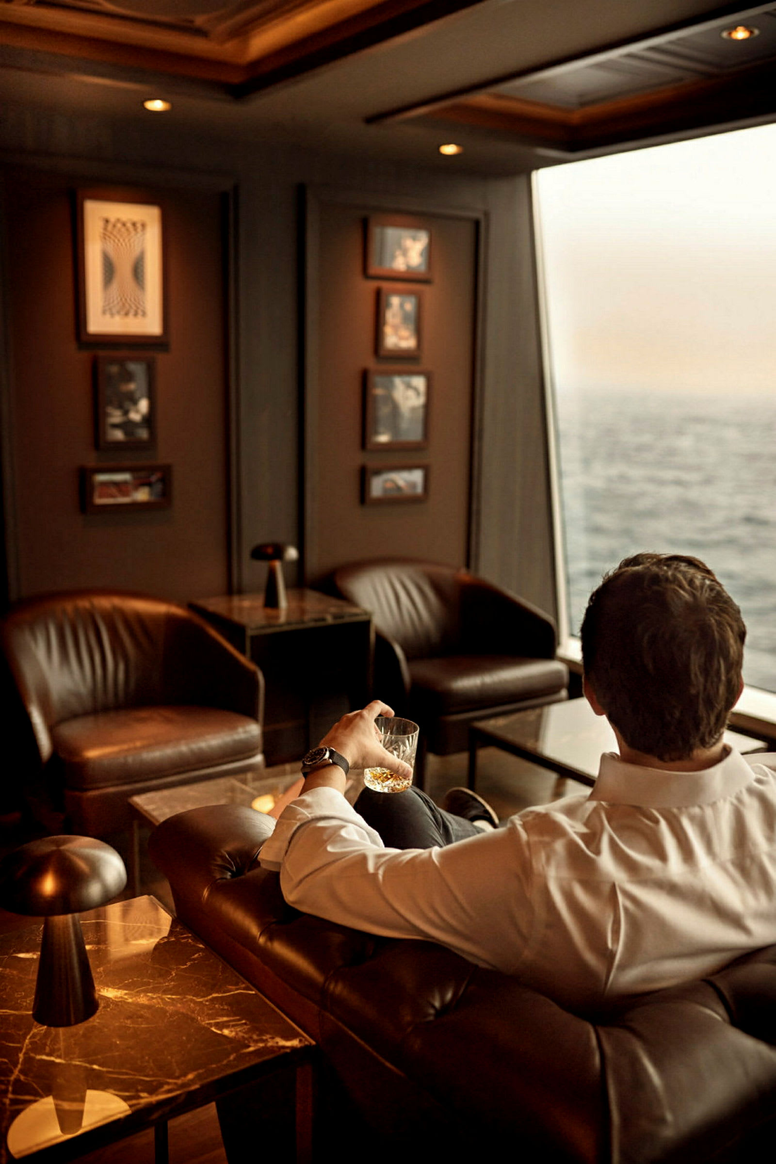 Man i vit skjorta sitter i en lounge med utsikt över havet och håller ett glas. (AI-genererad text)