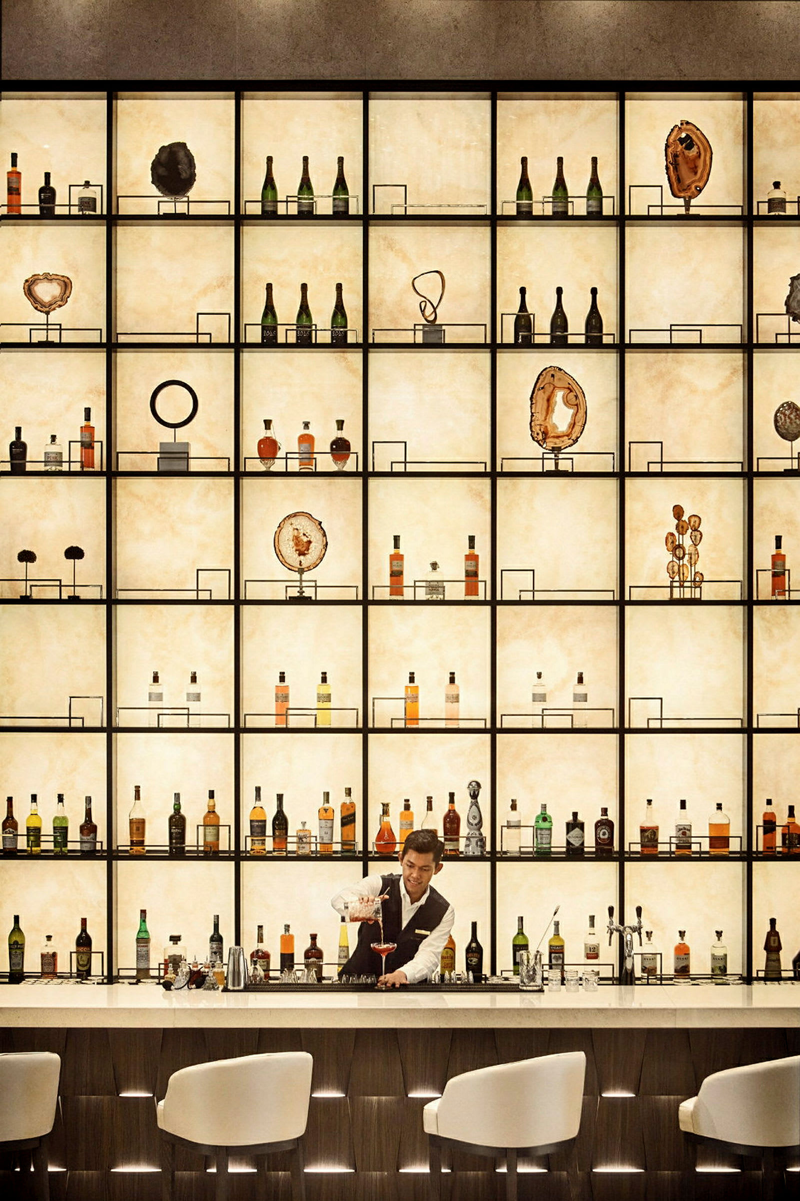 En bartendare serverar en drink vid en upplyst bokhylla fylld med flaskor och prydnadsföremål. (AI-genererad text)