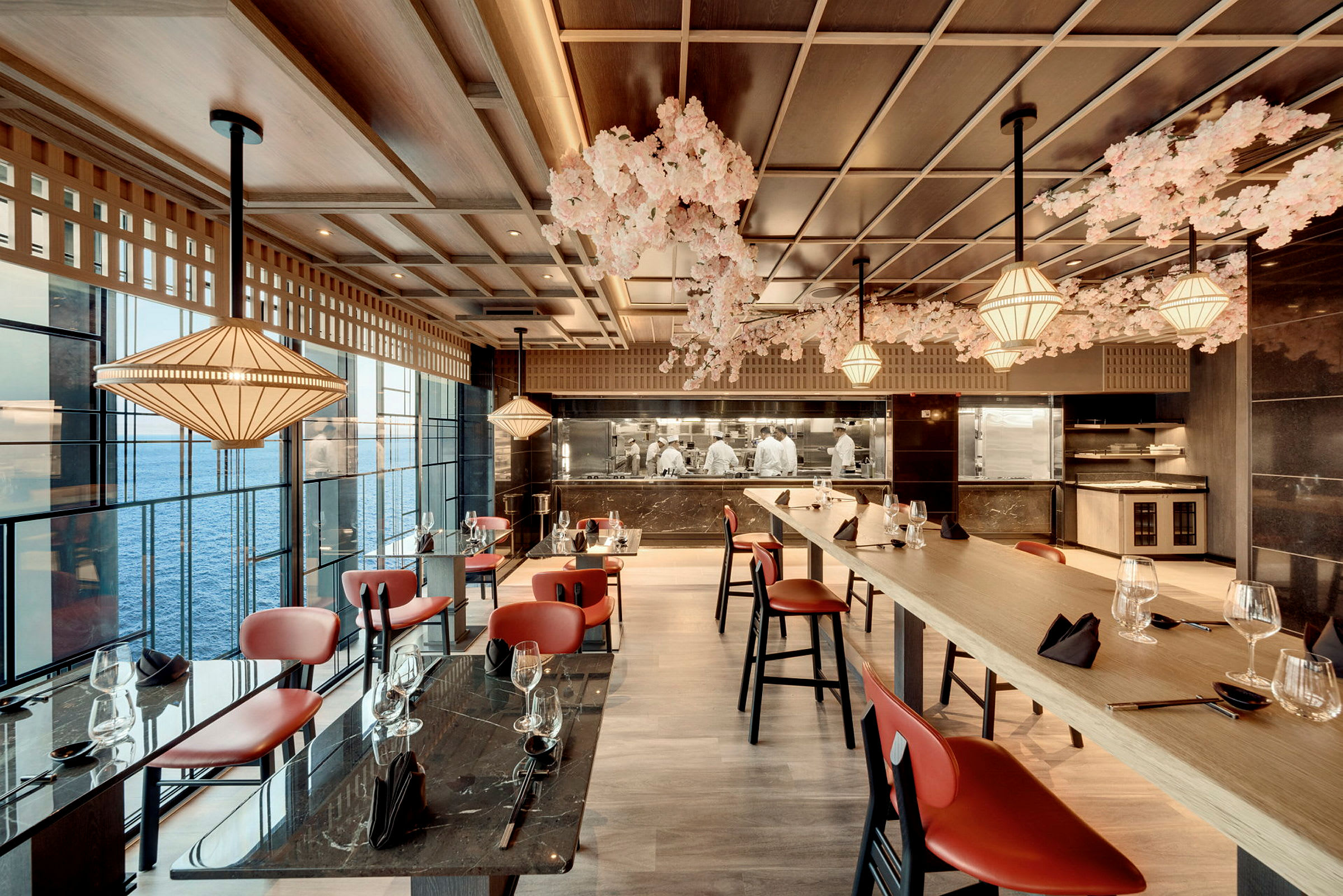 Modernt inredd restaurang med röda stolar, dekorationer i trä och glas, och en havsutsikt utanför fönstren. (AI-genererad text)