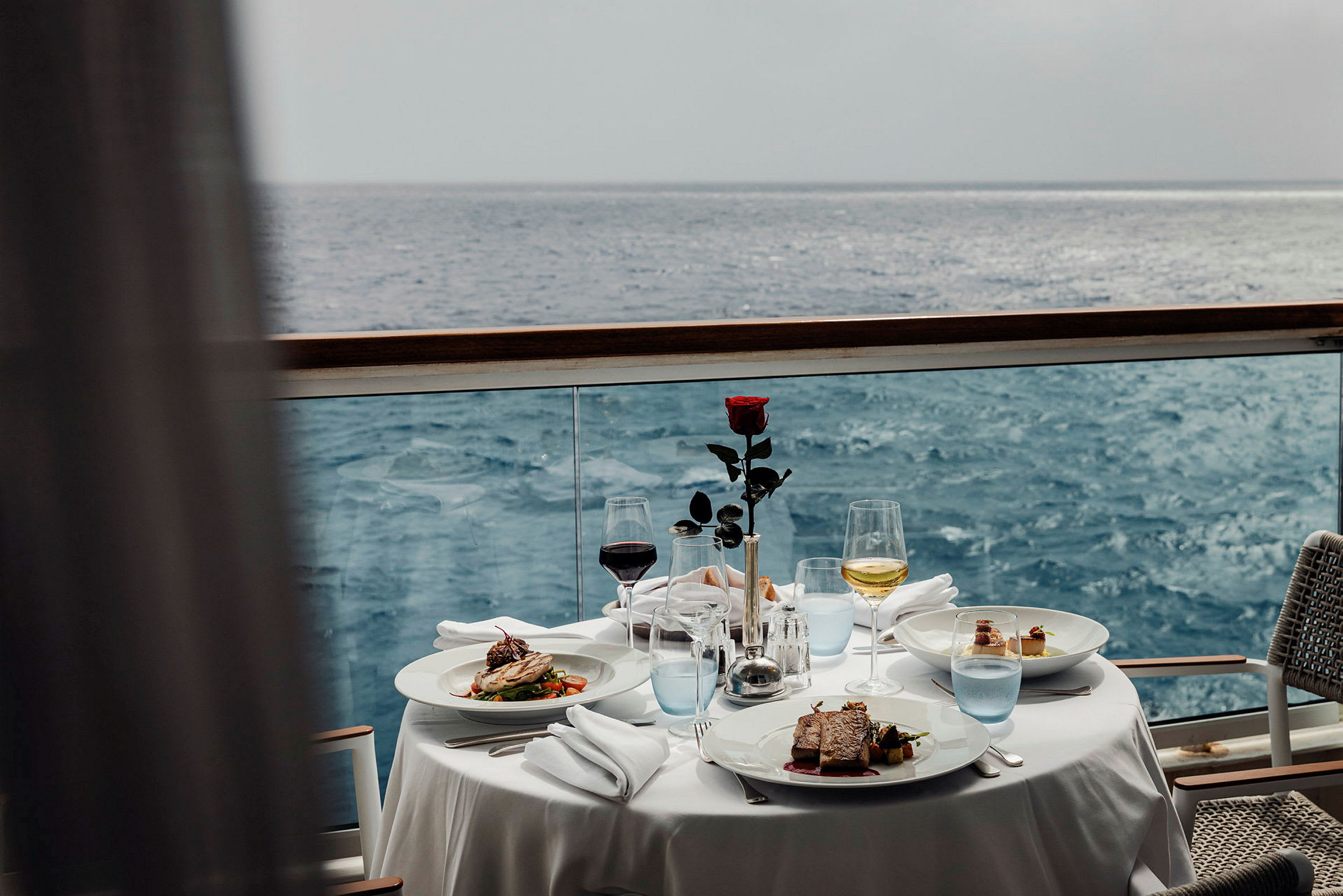 Ett bord med en vit duk, två tallrikar med mat och olika glas bredvid. Bordet står vid en balkong med utsikt över havet. (AI-genererad text)