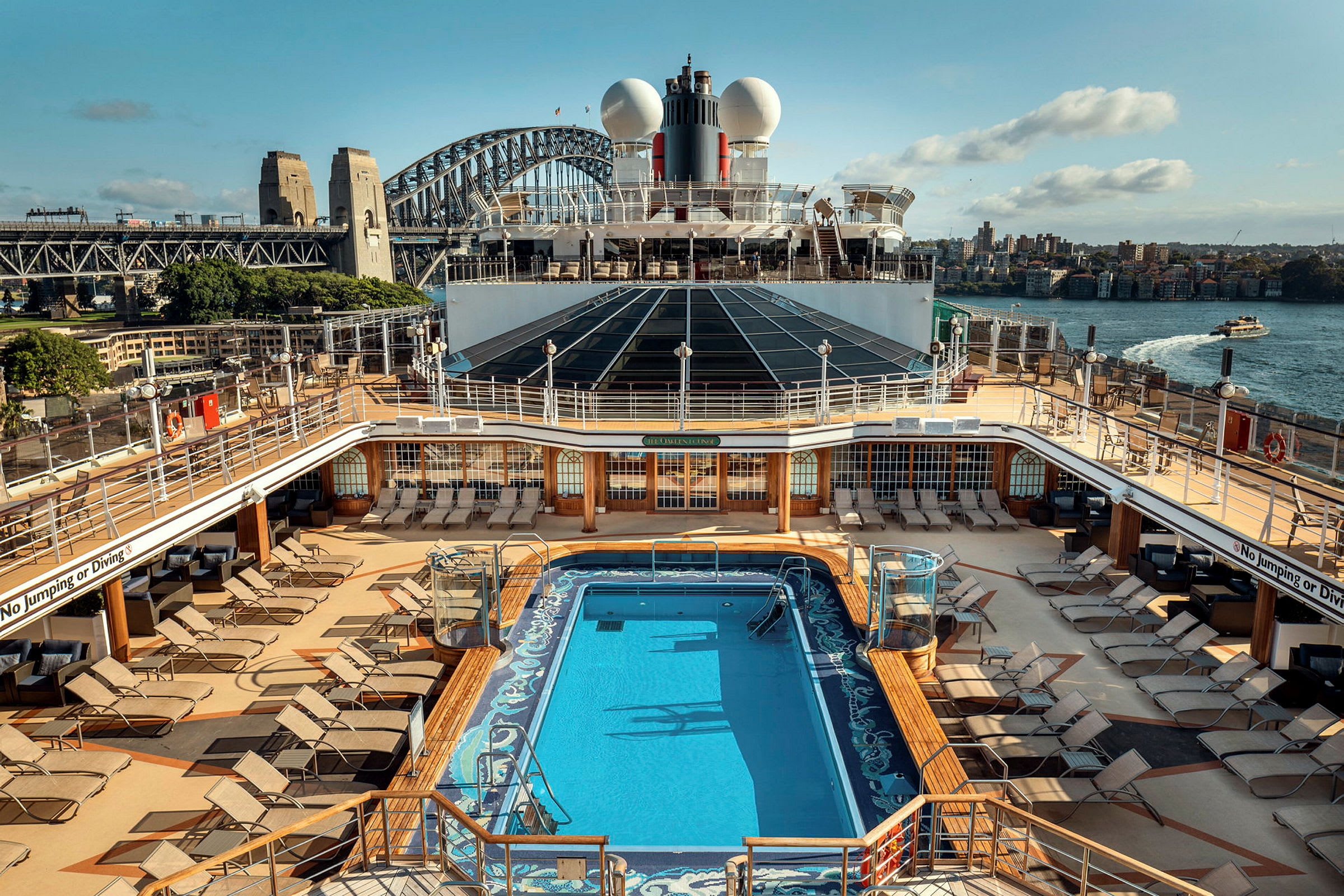 Bild på däcket av ett kryssningsfartyg med en pool och solstolar, Sydney Harbour Bridge i bakgrunden. (AI-genererad text)