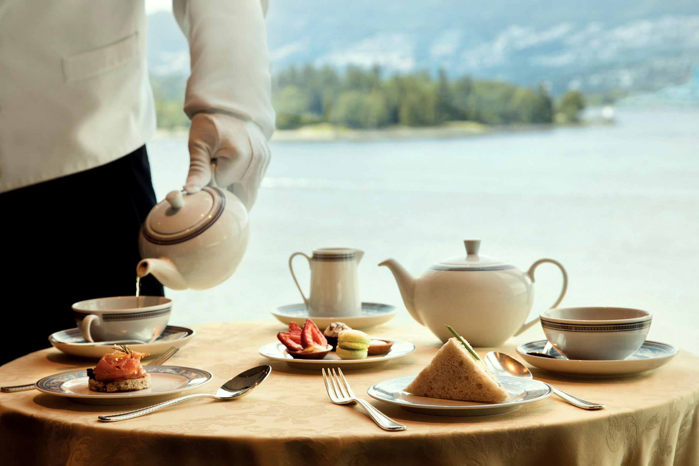 En servitör häller te från en kanna i en kopp vid ett bord med te och tillbehör. (AI-genererad text)