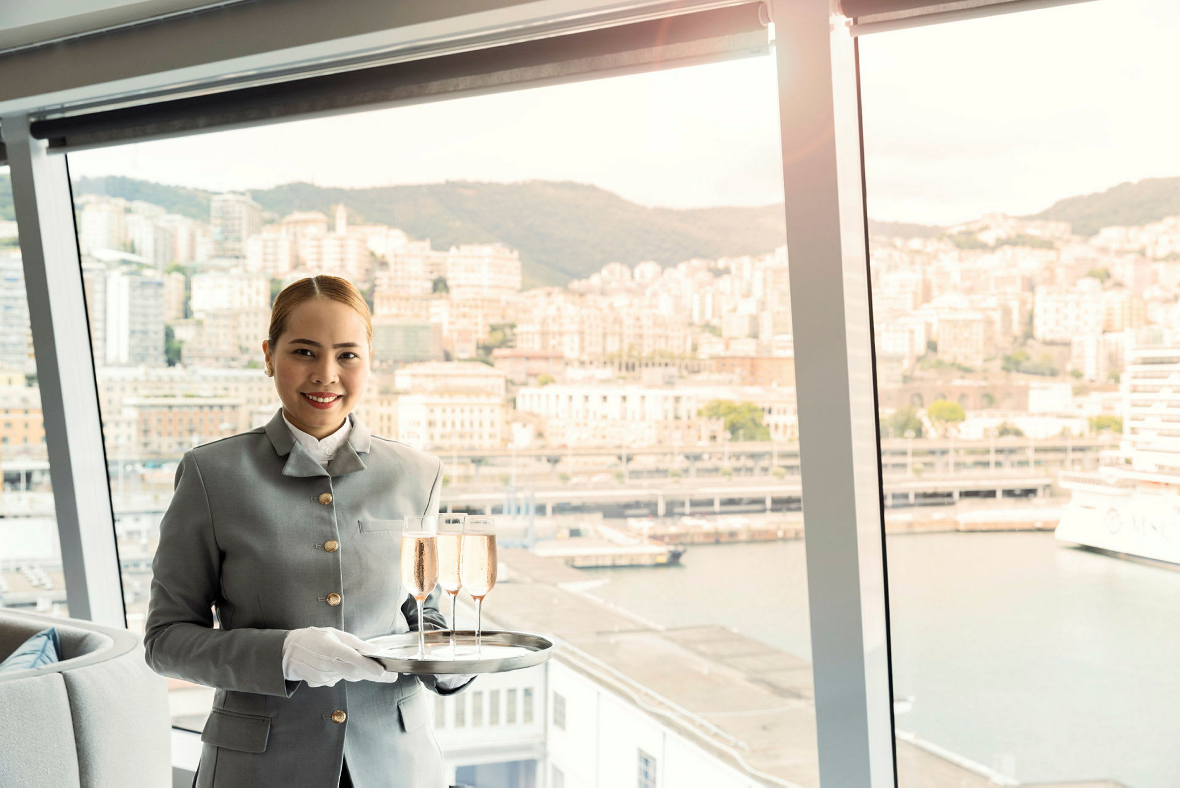 En person iklädd uniform håller en bricka med två glas champagne framför ett panoramafönster med utsikt över en stad och hamn. (AI-genererad text)