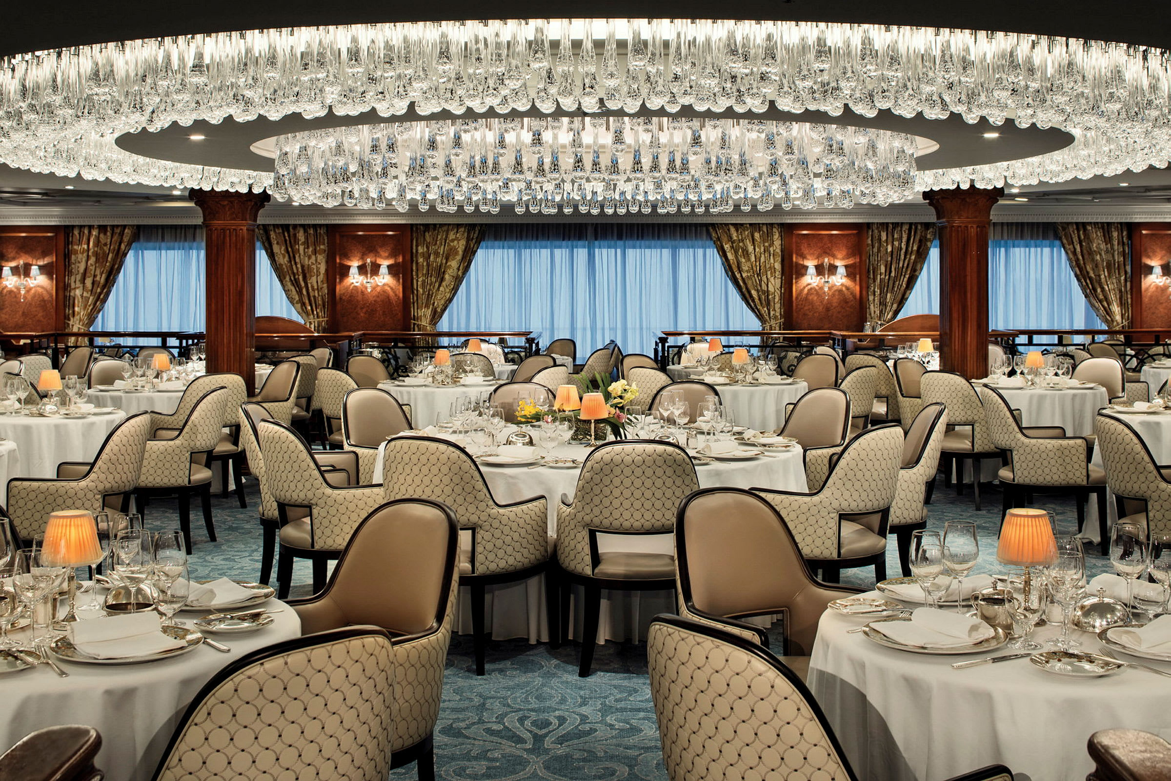 Interiör av en elegant restaurang med dukade bord, stolar, kristallkrona och gardiner framför fönster. (AI-genererad text)
