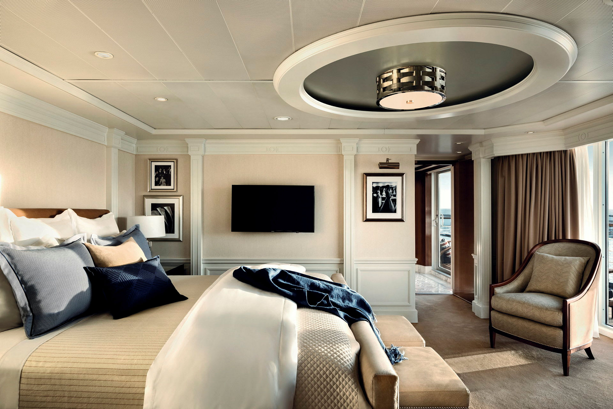 Ett elegant hotellrum med en stor säng, sittmöbler, en väggmonterad TV, och stora fönster som släpper in dagsljus. (AI-genererad text)