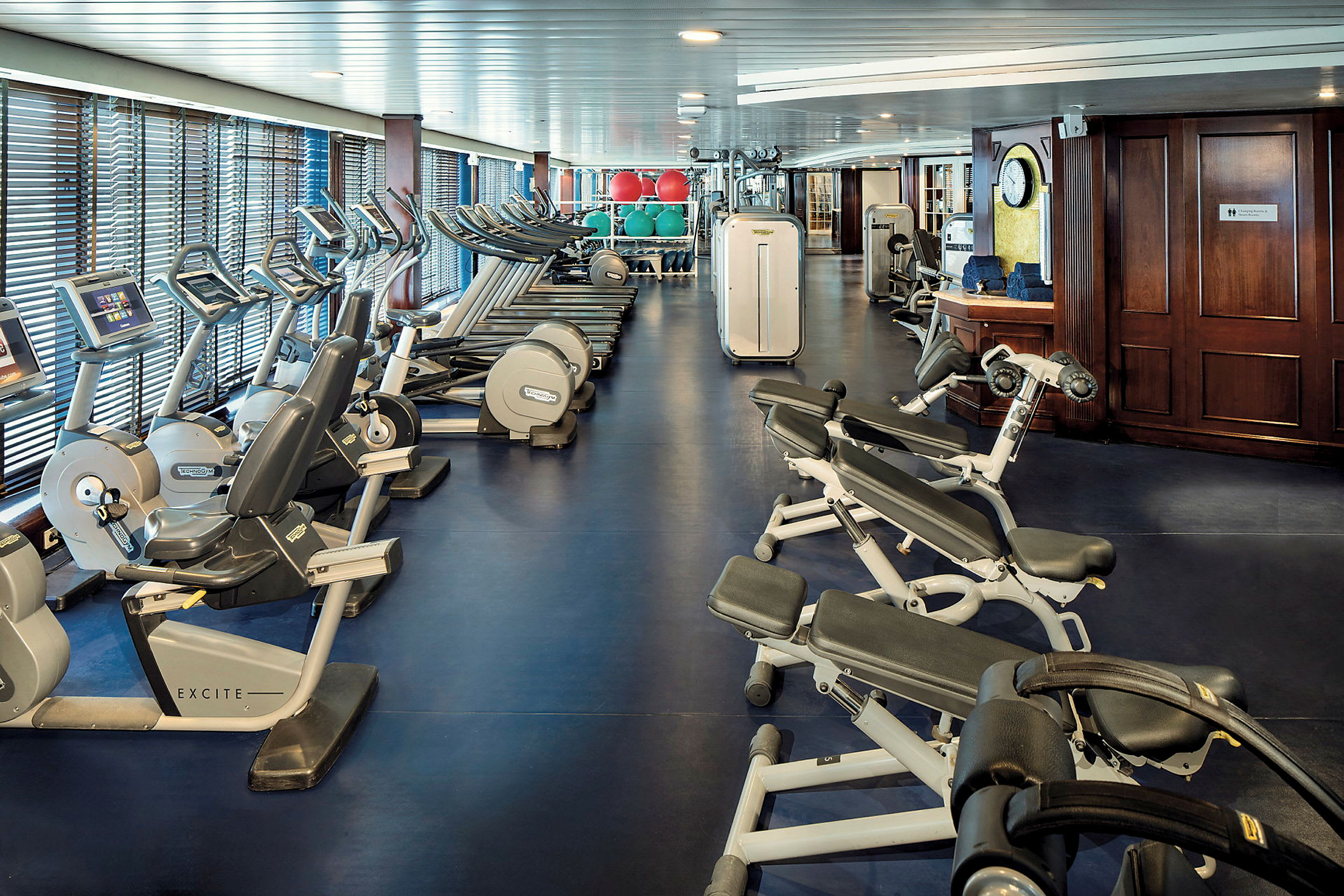 Ett gym med träningsmaskiner och styrketräningsutrustning. Fönster linjerar ena sidan av rummet. (AI-genererad text)