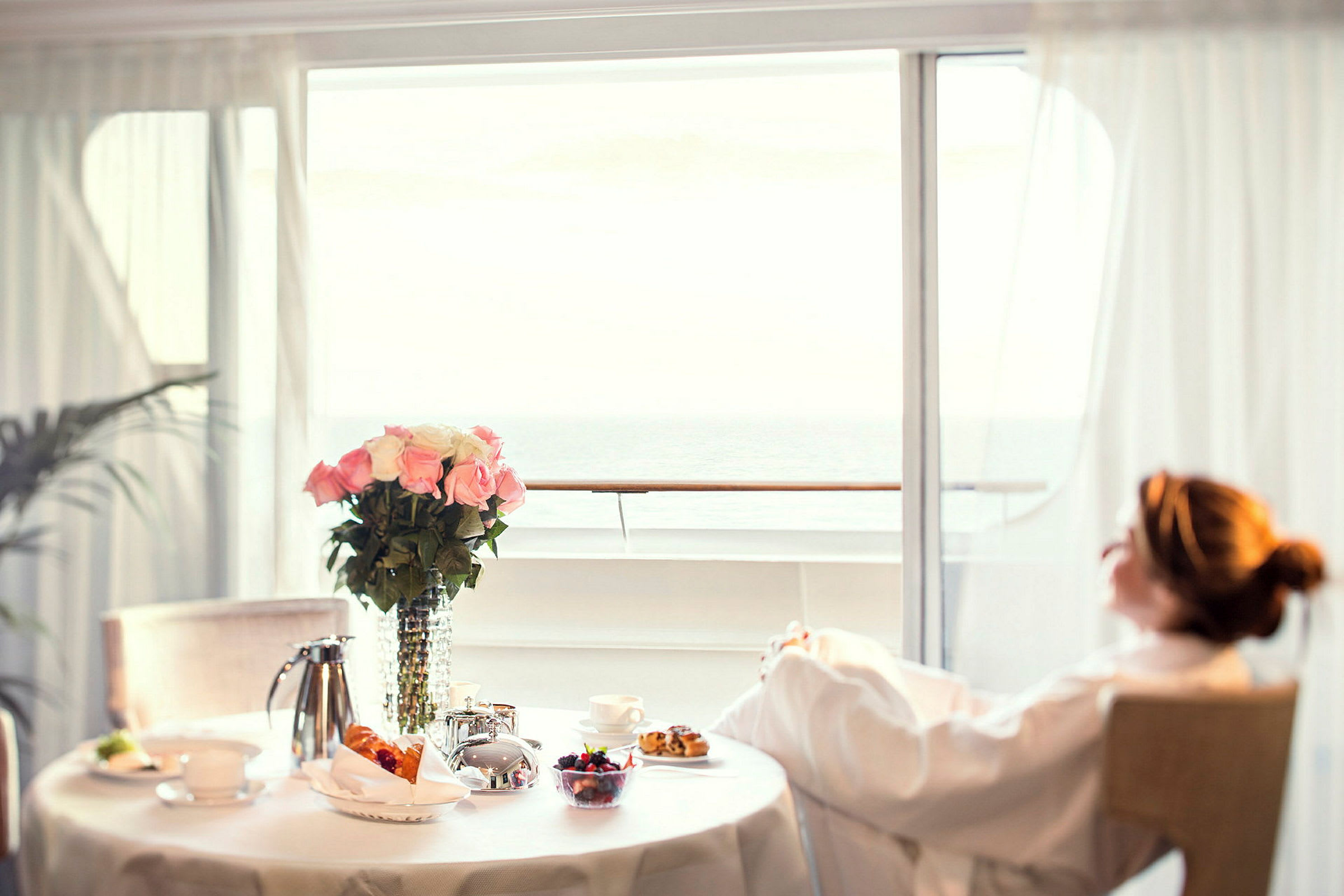 En person sitter avslappnad i en stol vid ett dukat bord nära ett stort fönster med utsikt över havet. På bordet finns blommor, te och frukost. (AI-genererad text)