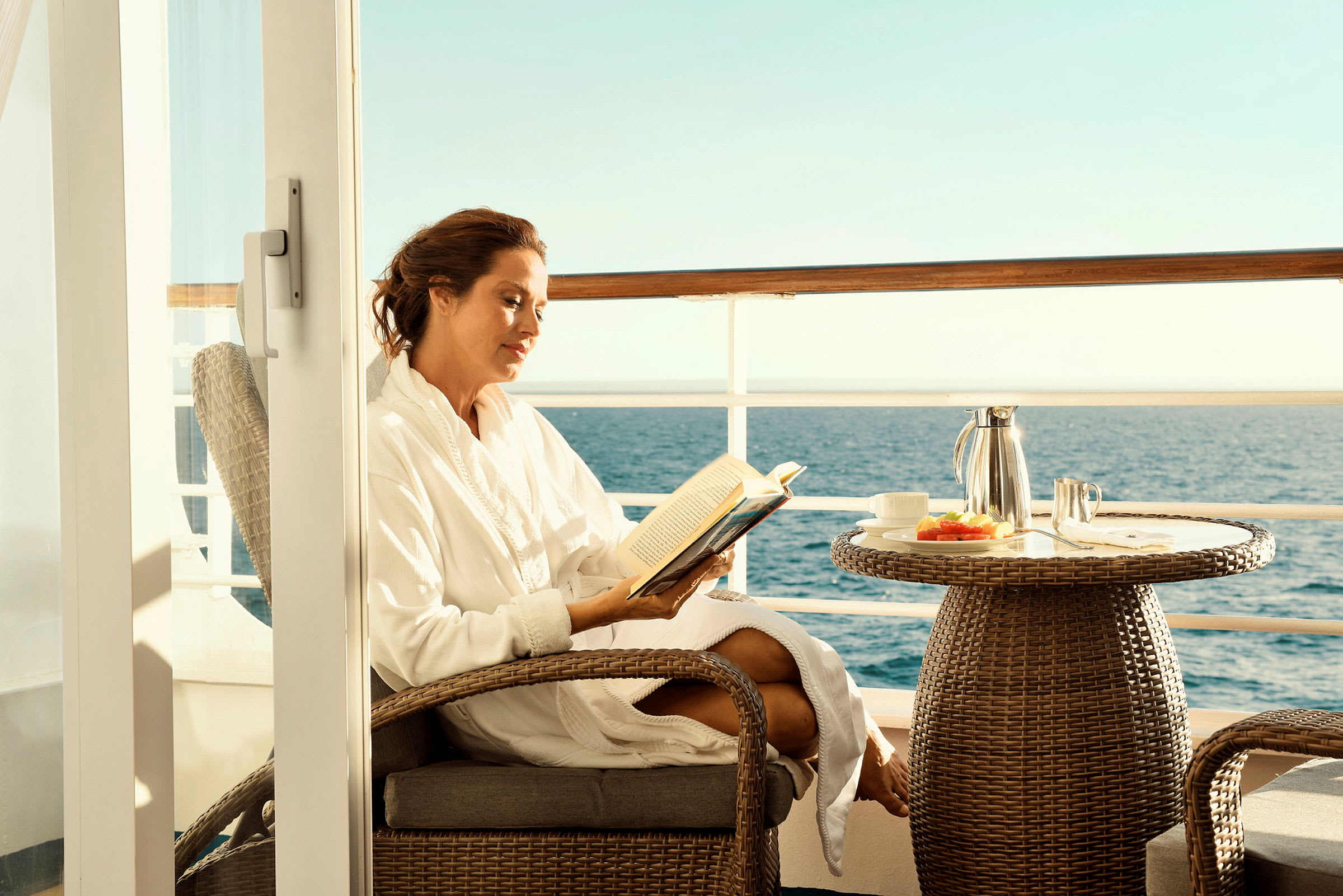 Person sitter bekvämt på en balkong med utsikt över havet och läser en bok, bredvid ett bord med serverad frukost. (AI-genererad text)