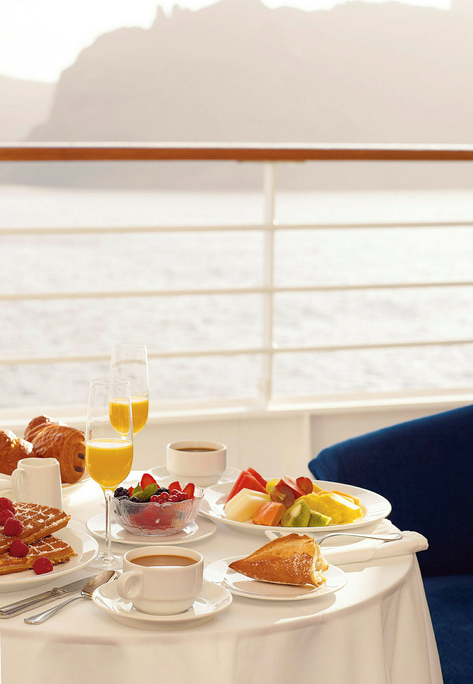 Frukostservering på ett bord med en vy över vattnet. Bordet innehåller bland annat frukt, bröd, juicelglas och kaffe. (AI-genererad text)