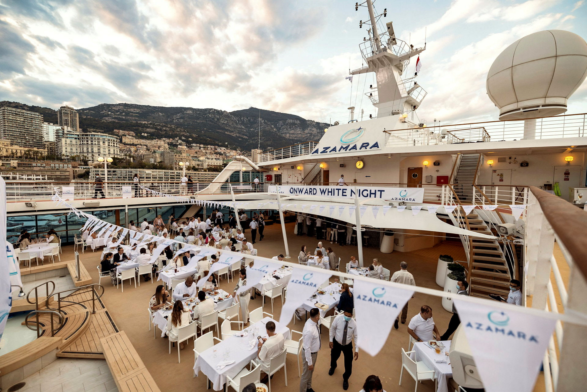 Utomhusmiljö på ett kryssningsfartyg med uppställda bord och stolar för en middag, omgiven av flaggor med text och logotyp för Azamara. (AI-genererad text)