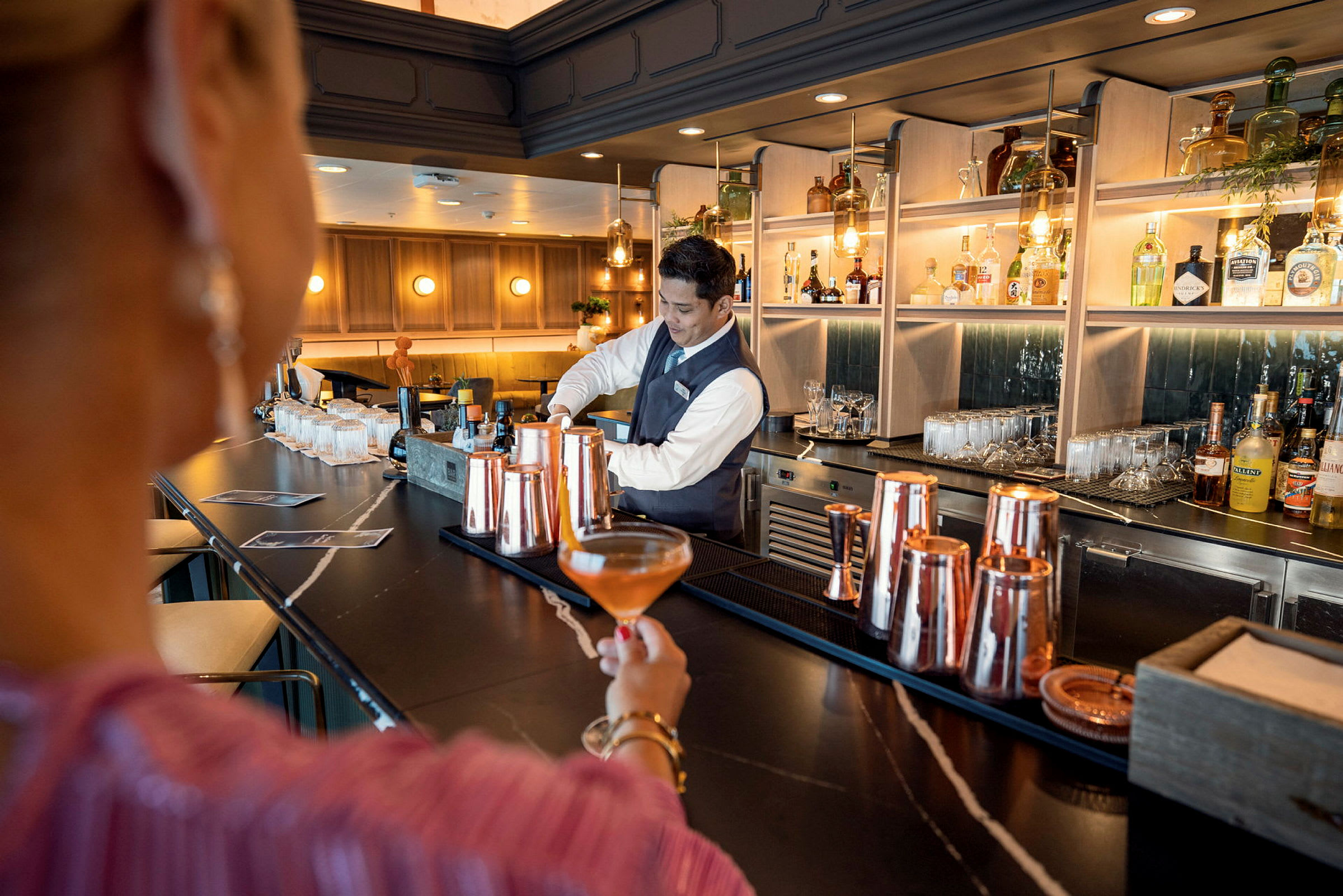 En person håller en drink vid en elegant bardisk, där en bartender serverar. (AI-genererad text)