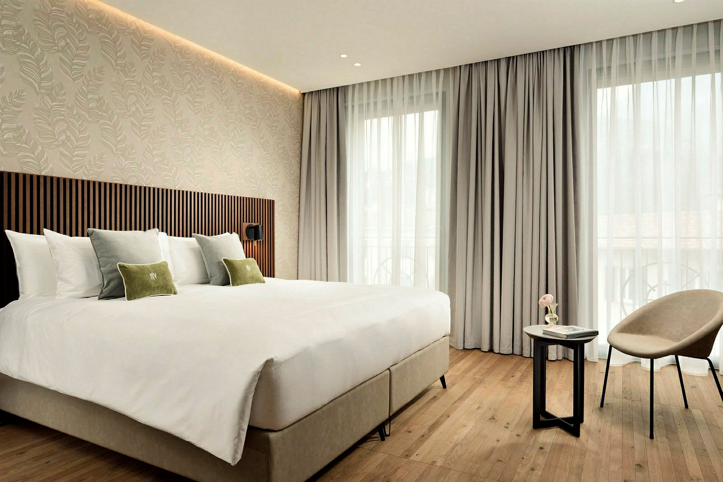 Ett modernt hotellrum med en stor säng, dekorativa kuddar, en fåtölj och ett mindre bord framför en fönstervägg med ljusa gardiner. (AI-genererad text)