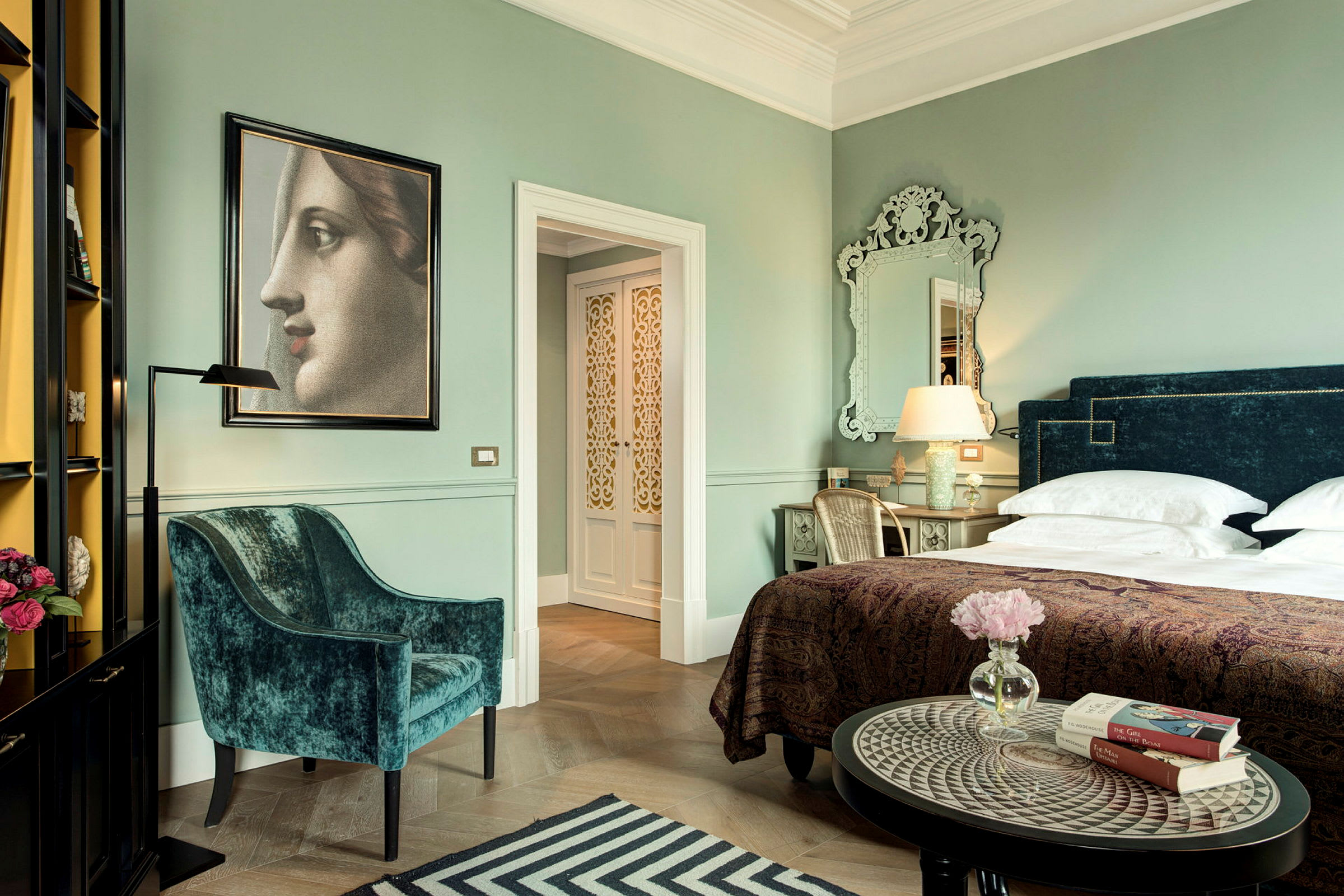 En elegant hotellsvit med grön vägg, en mjuk sammetsstol, en säng med mörkblå sänggavel och prydnadsdetaljer som bordslampor och blommor. (AI-genererad text)