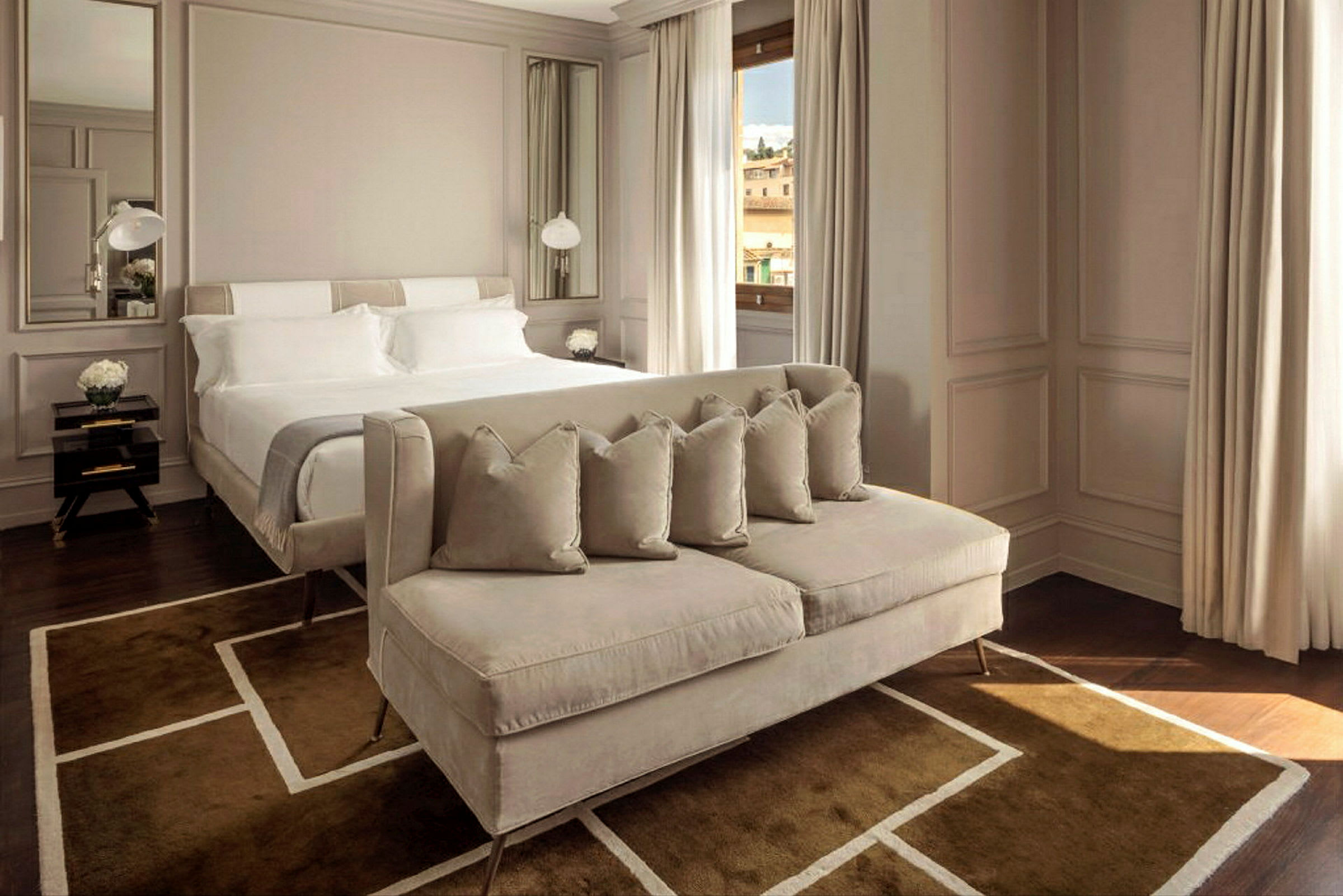 Ett elegant hotellrum med en bred säng och en sittmöbel framför. Rummet är inrett i ljusa toner och har stora fönster. (AI-genererad text)