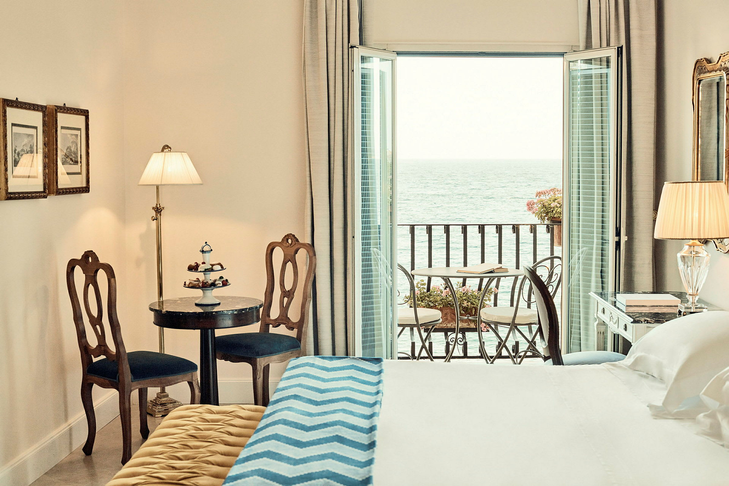Ett hotellrum med en säng, bord med stolar och en balkong med utsikt över havet. (AI-genererad text)