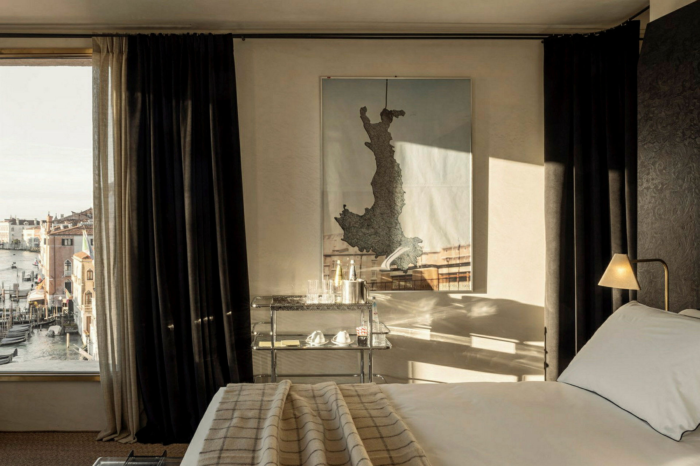 Ett hotellrum med en säng, ett nattduksbord och en karta på väggen, samt ett fönster med utsikt över gondoler och byggnader. (AI-genererad text)