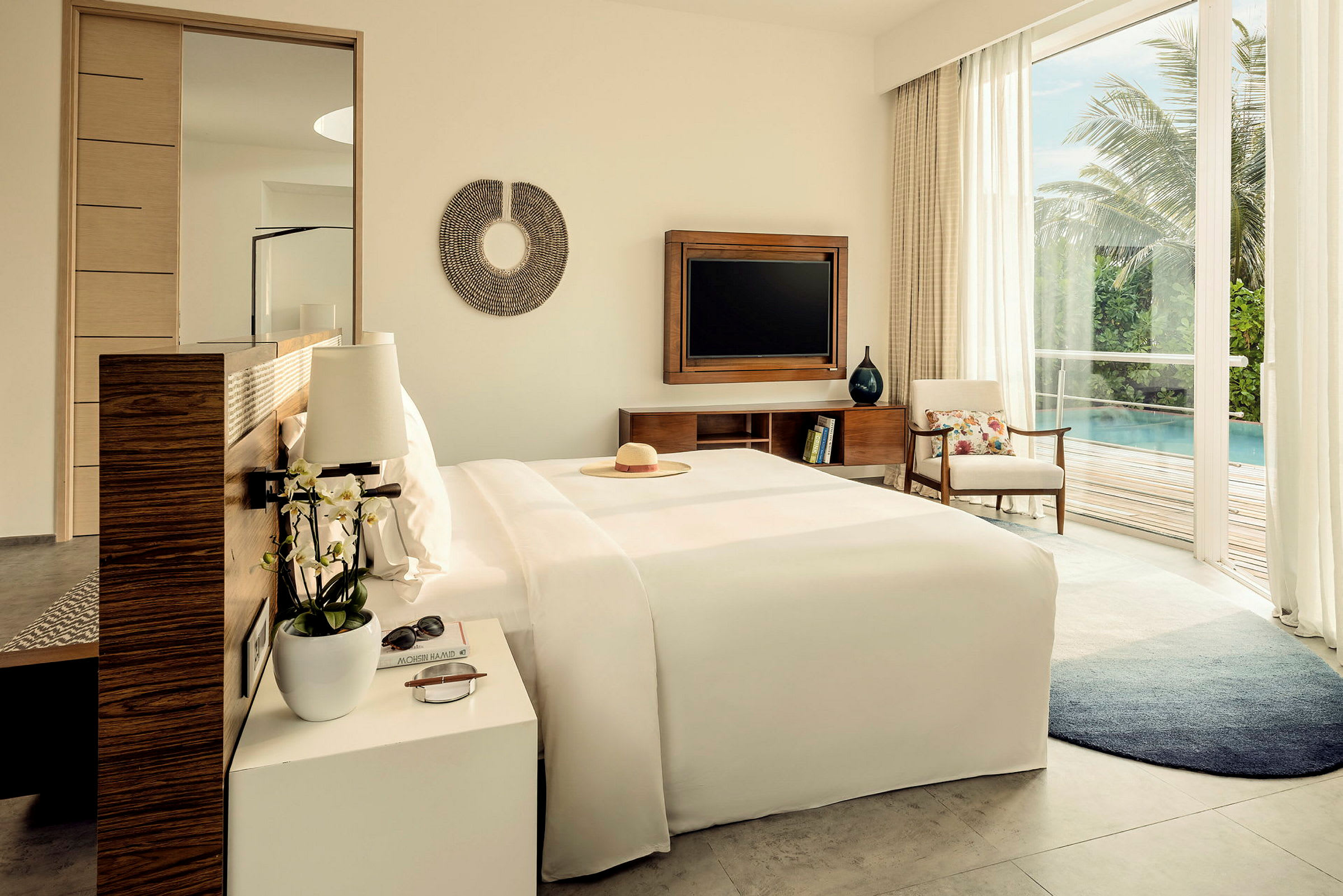 Ett hotellrum med stor säng, sidobord och TV. Utsikt mot en terrass med pool och grönska. (AI-genererad text)
