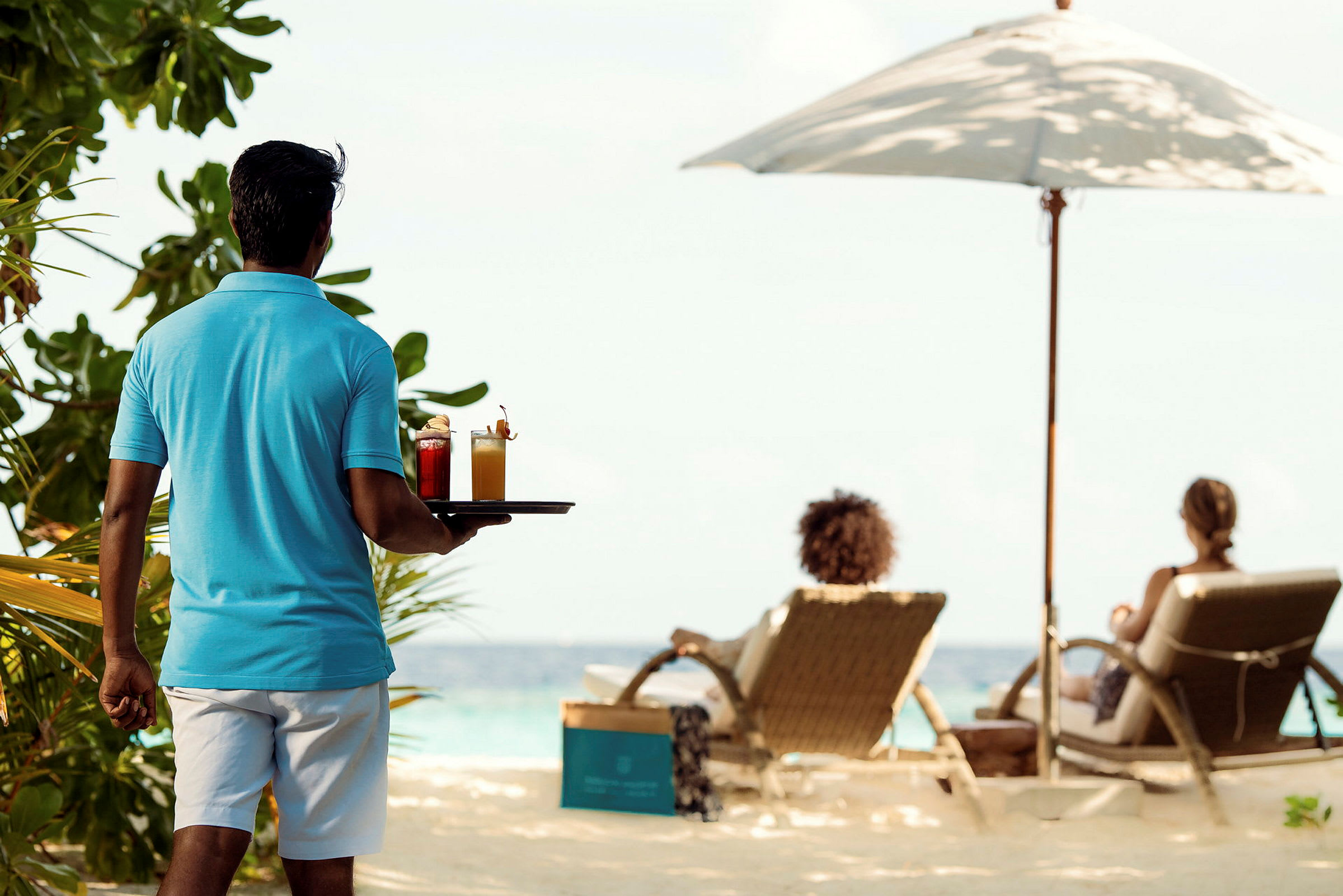 En person går mot en strand och håller en bricka med två cocktails. Två personer sitter och kopplar av på solstolar under ett parasoll vid stranden. (AI-genererad text)