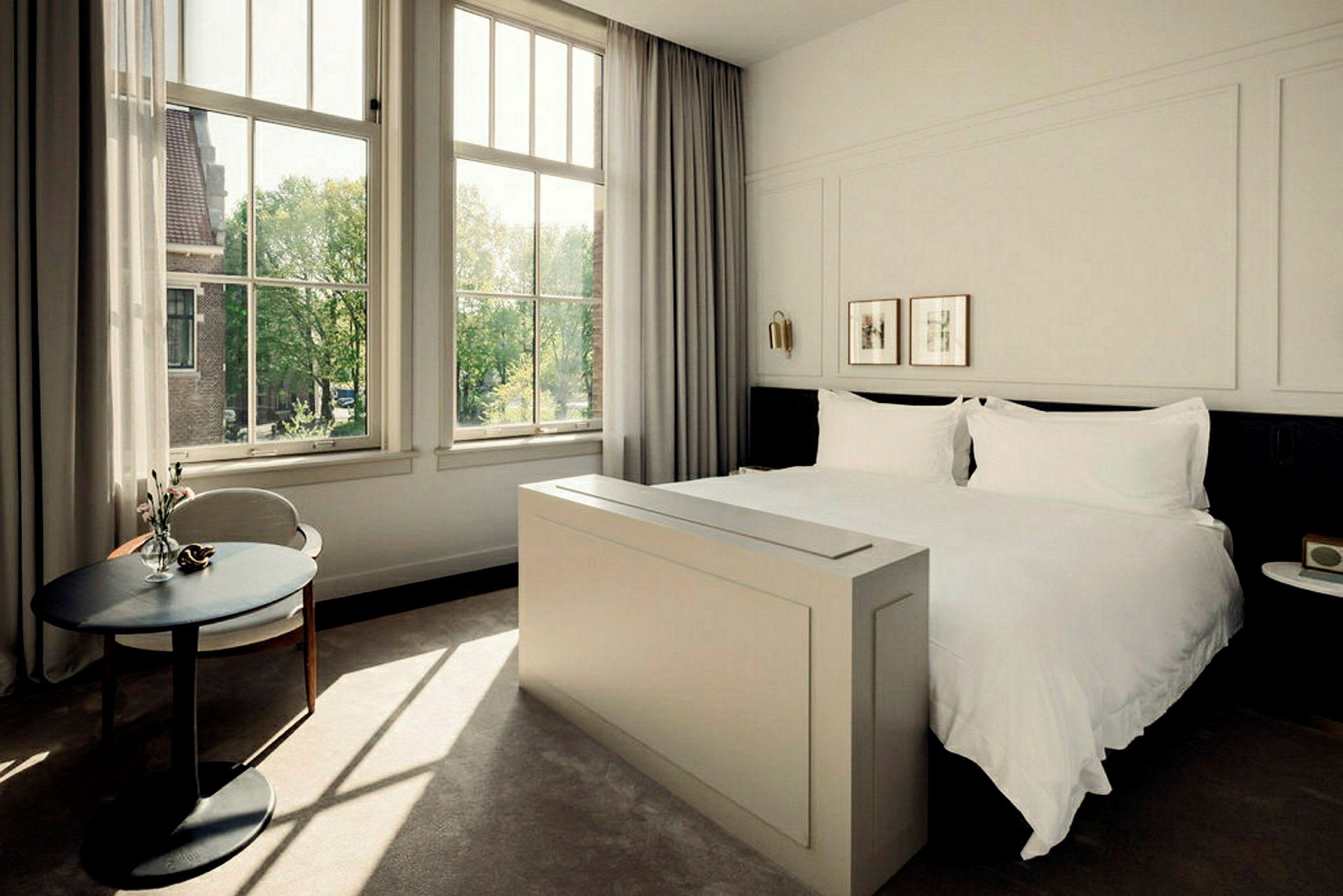 Ett ljust och elegant hotellrum med en stor säng, stora fönster och ett litet runt bord. (AI-genererad text)