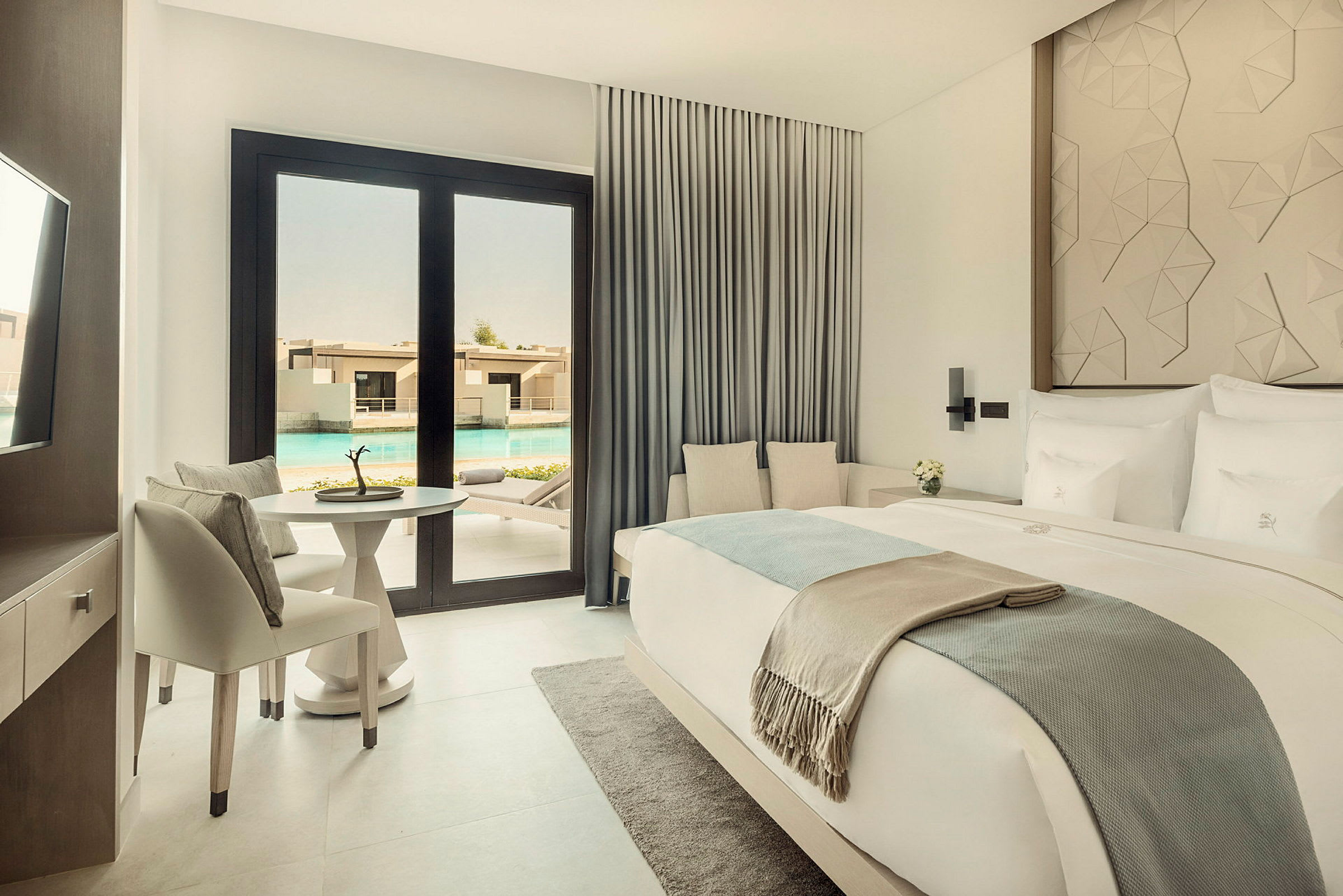 Ett elegant hotellrum med en dubbelsäng, sittgrupp, bord och stolar, med utsikt över en pool genom glasdörrar. (AI-genererad text)