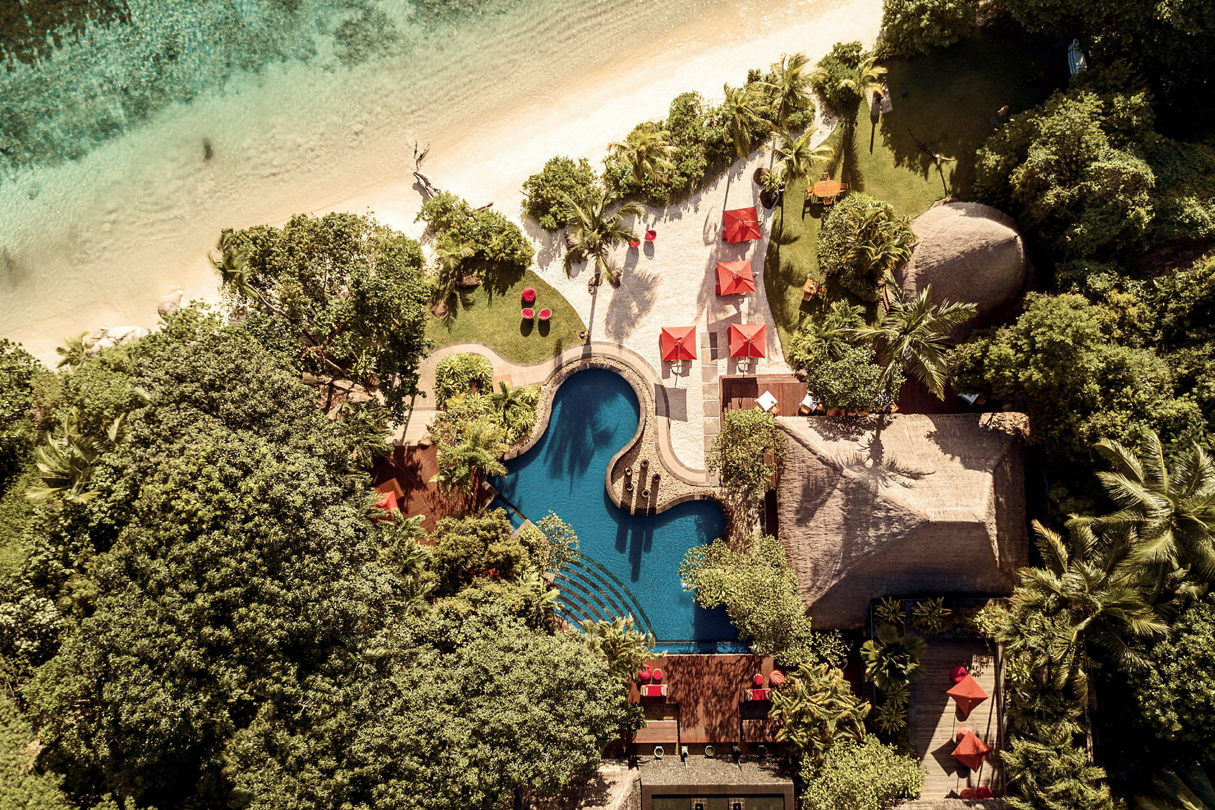 Flygfoto av resortområde med pool, omgivande vegetation, sandstrand och röda parasoller. (AI-genererad text)