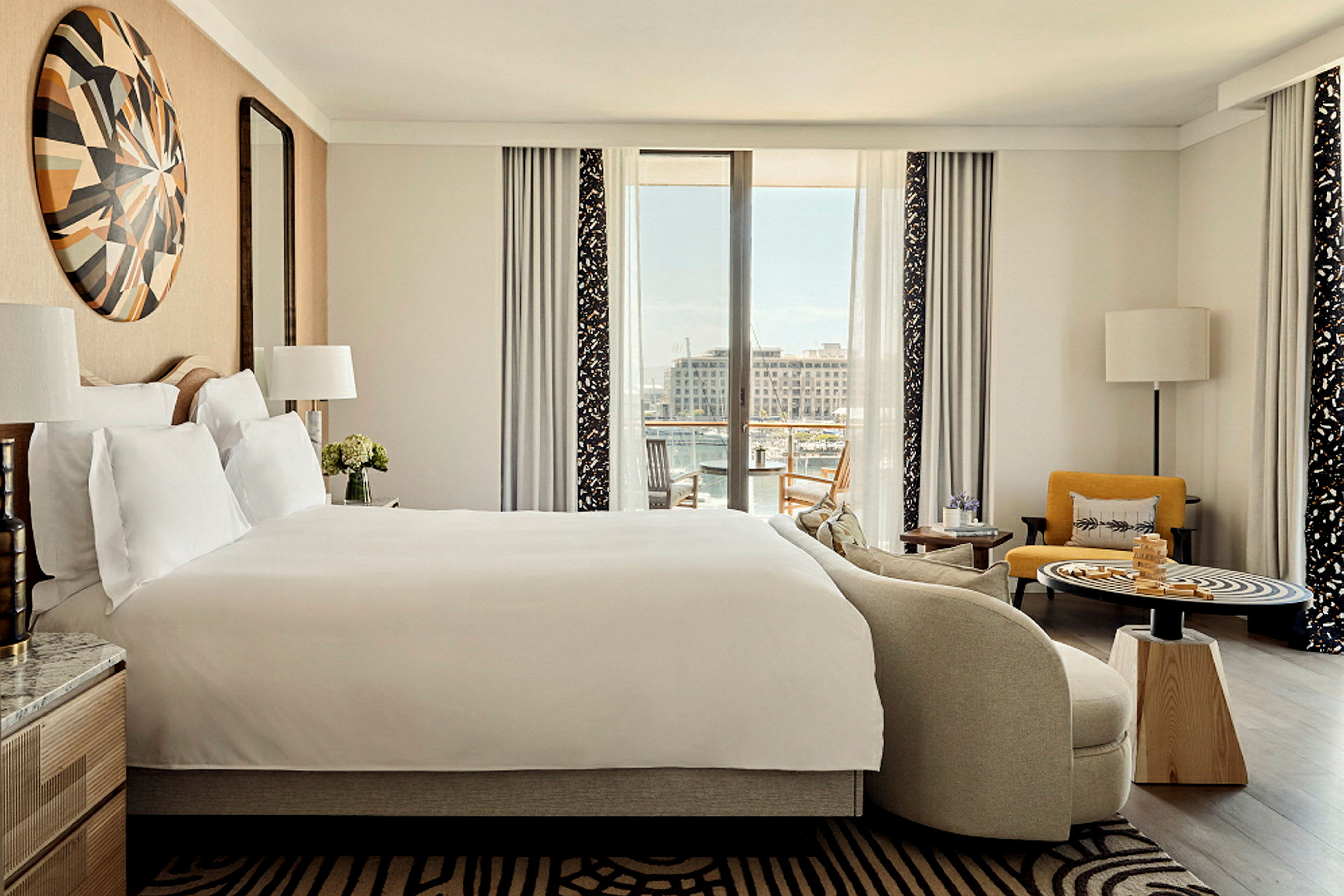 Ett hotellrum med stor säng, sittmöbler och en utsikt mot en marina genom stora fönster. (AI-genererad text)