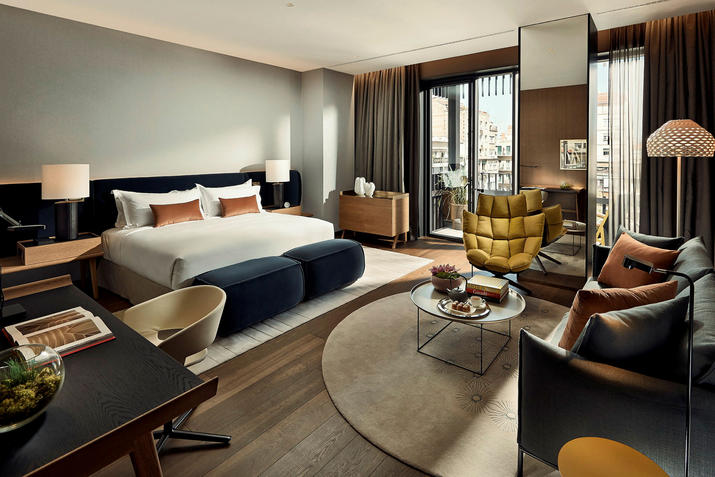 Ett modernt hotellrum med stor säng, sittgrupp och utsikt mot byggnader. (AI-genererad text)